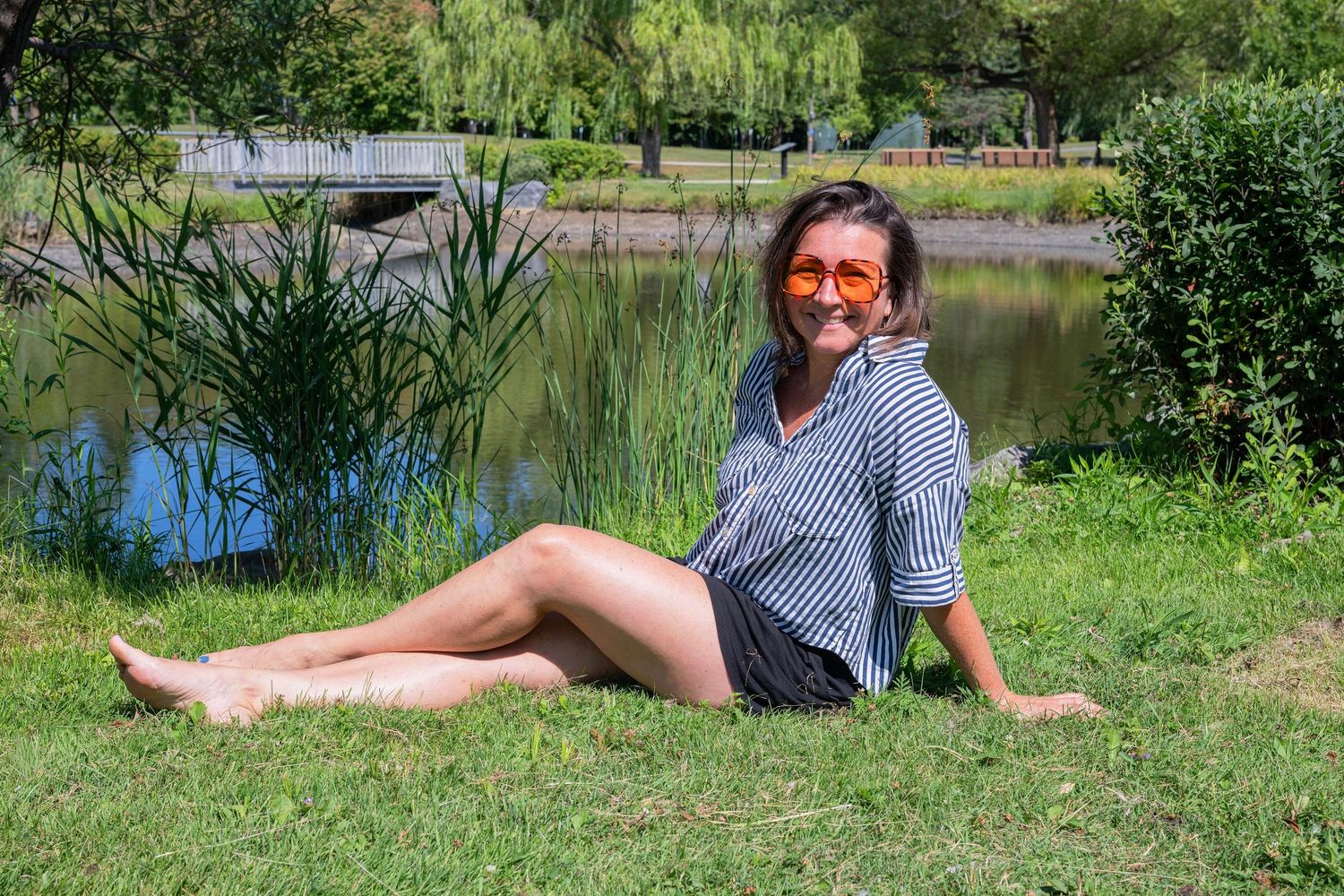 Smiling woman in striped shirt and shorts sitting barefoot on grass by a peaceful pond outdoors.