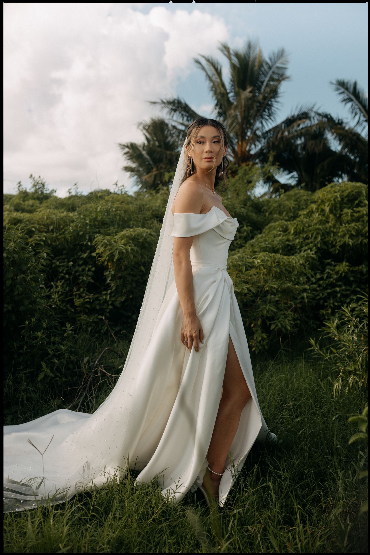 Bride in off-shoulder white gown with veil stands in lush tropical greenery, elegant wedding dress with thigh-high slit.