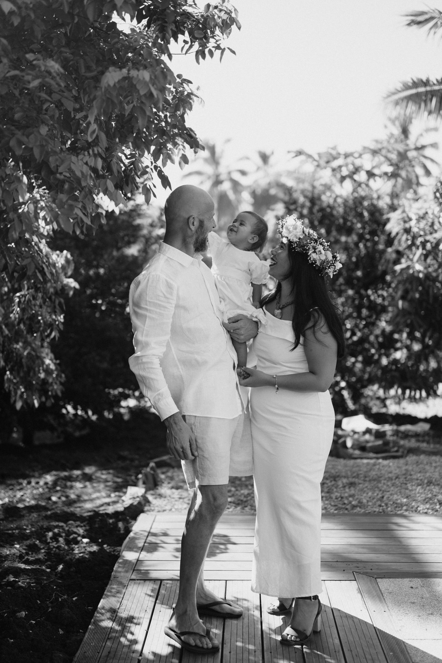 Black and white photo of a couple in white attire holding their baby on a wooden deck surrounded by tropical trees.