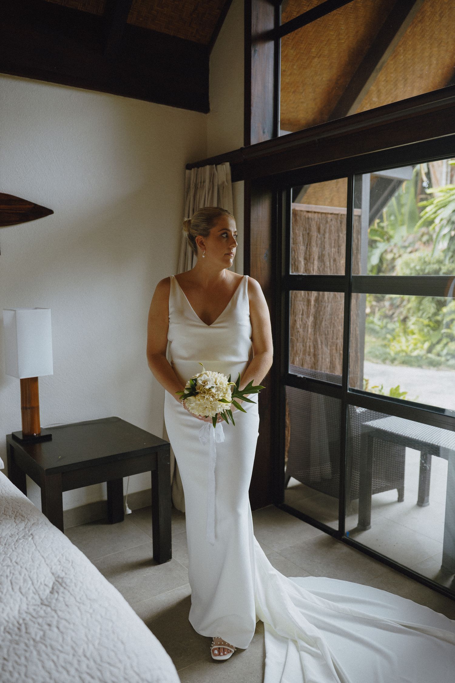 Bride in white satin gown holding bouquet stands by large window in tropical villa bedroom.