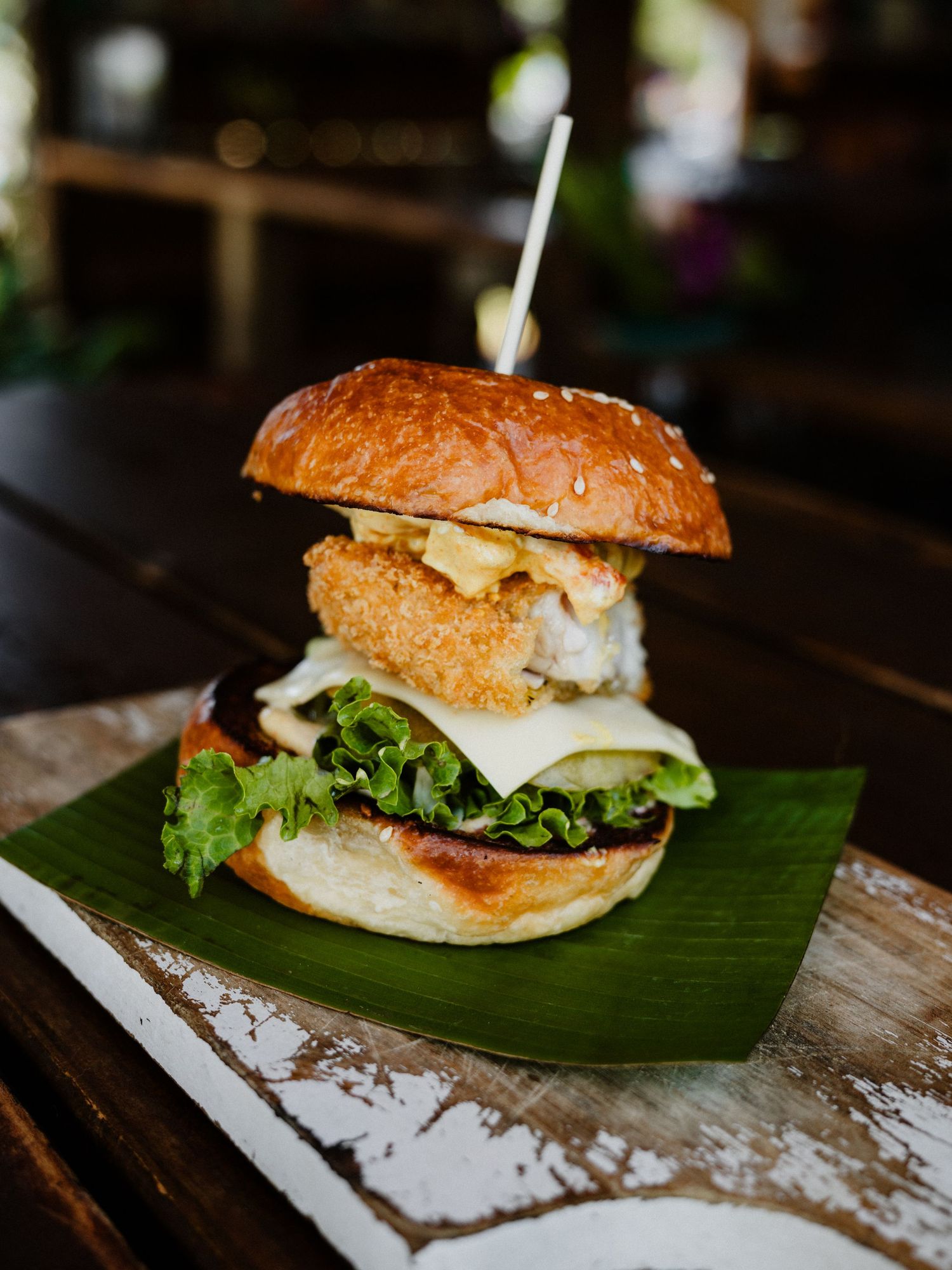 A gourmet crispy chicken burger with lettuce and cheese on a banana leaf, served on a rustic wooden table.
