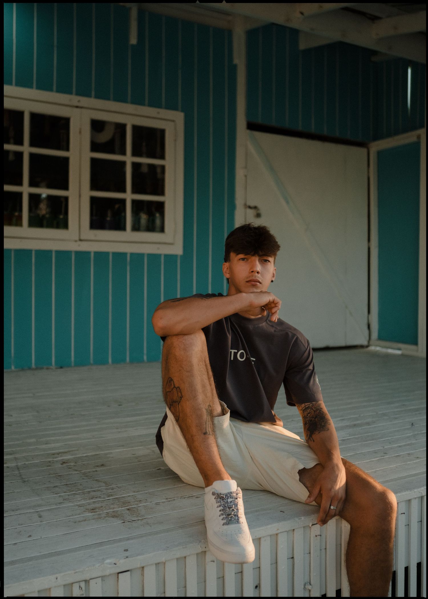 Young tattooed man in gray t-shirt and cream pants sitting on white porch of teal building.