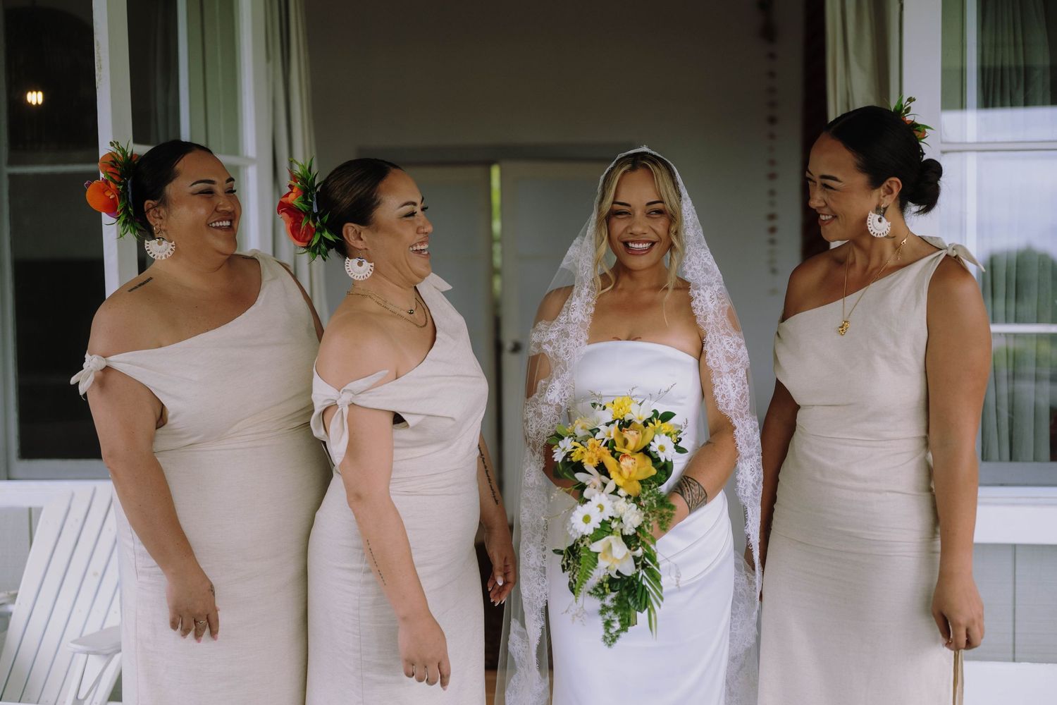 Bride in white gown holding yellow bouquet poses with three bridesmaids in ivory dresses on a porch.