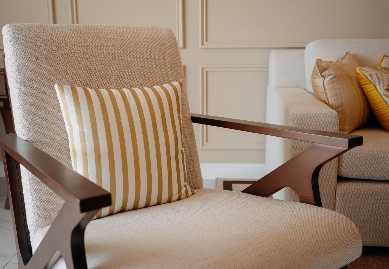 Beige mid-century modern armchair with wood arms featuring a gold striped accent pillow in an elegant neutral living room.