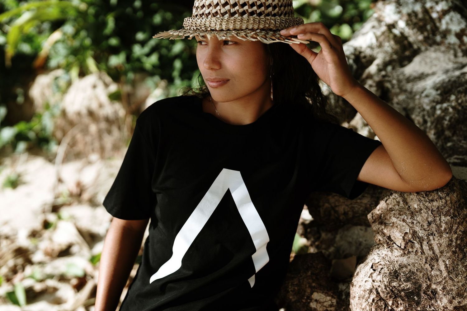 Young person wearing a black graphic tee with triangle logo and woven straw hat, posing outdoors near rocks.