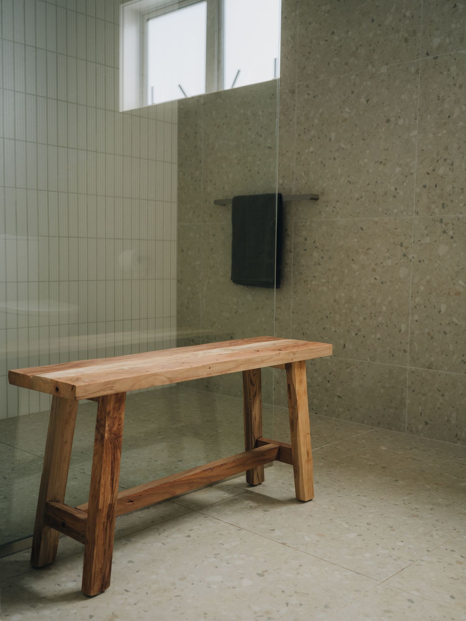 Rustic wooden bench in a modern bathroom with terrazzo floors, white tiles, and a dark towel hanging on the wall.