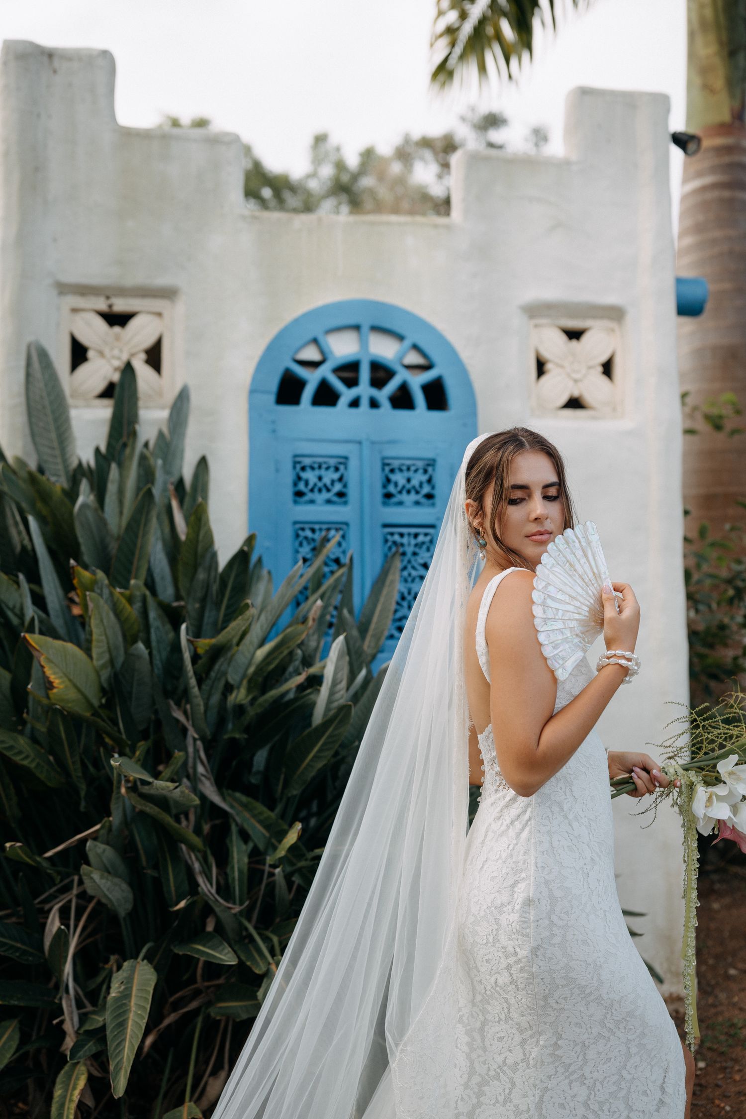 Bride in lace gown holds fan veil before blue Moroccan doors and tropical greenery.