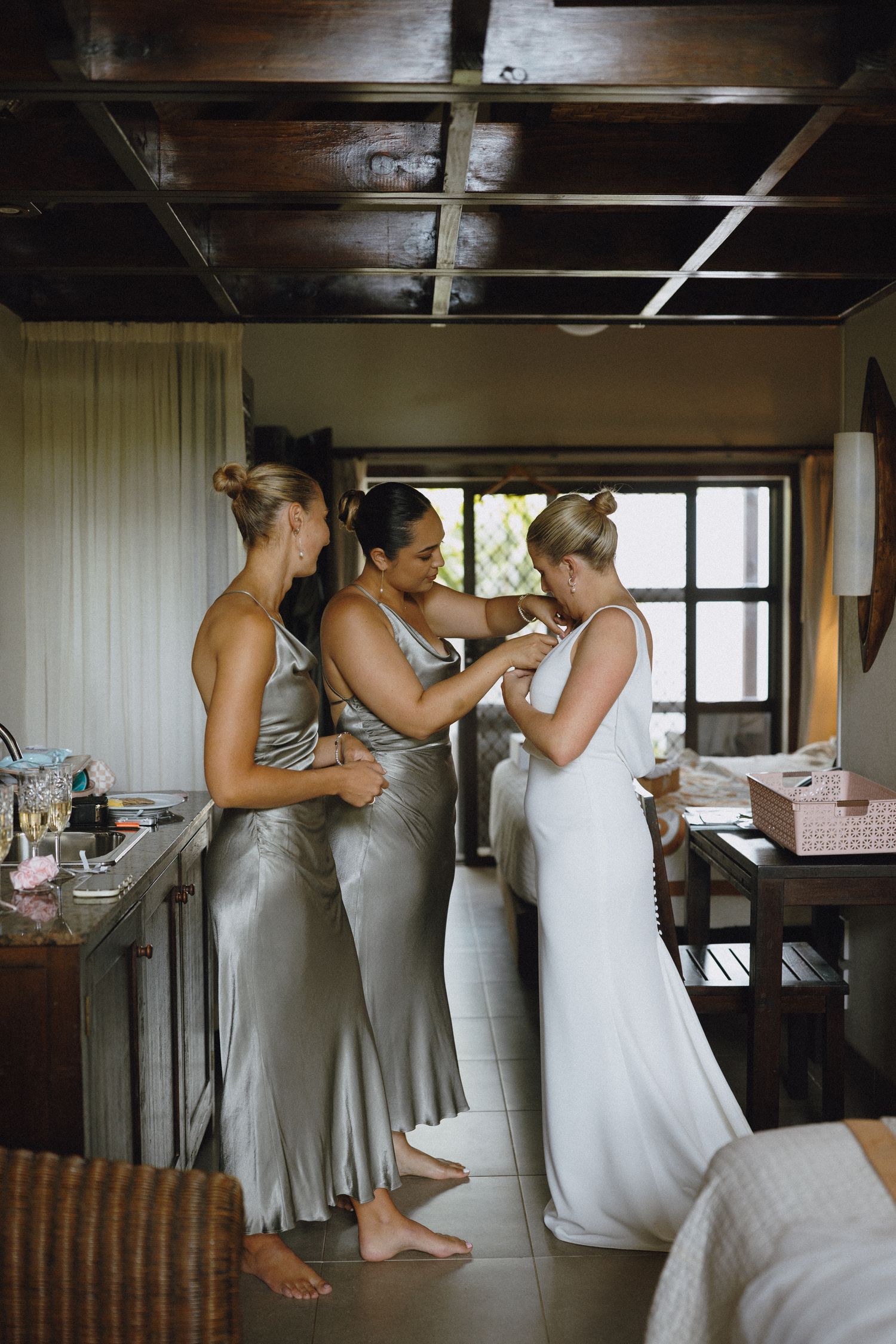 Bride in white gown assisted by two bridesmaids in silver dresses during wedding preparations indoors.