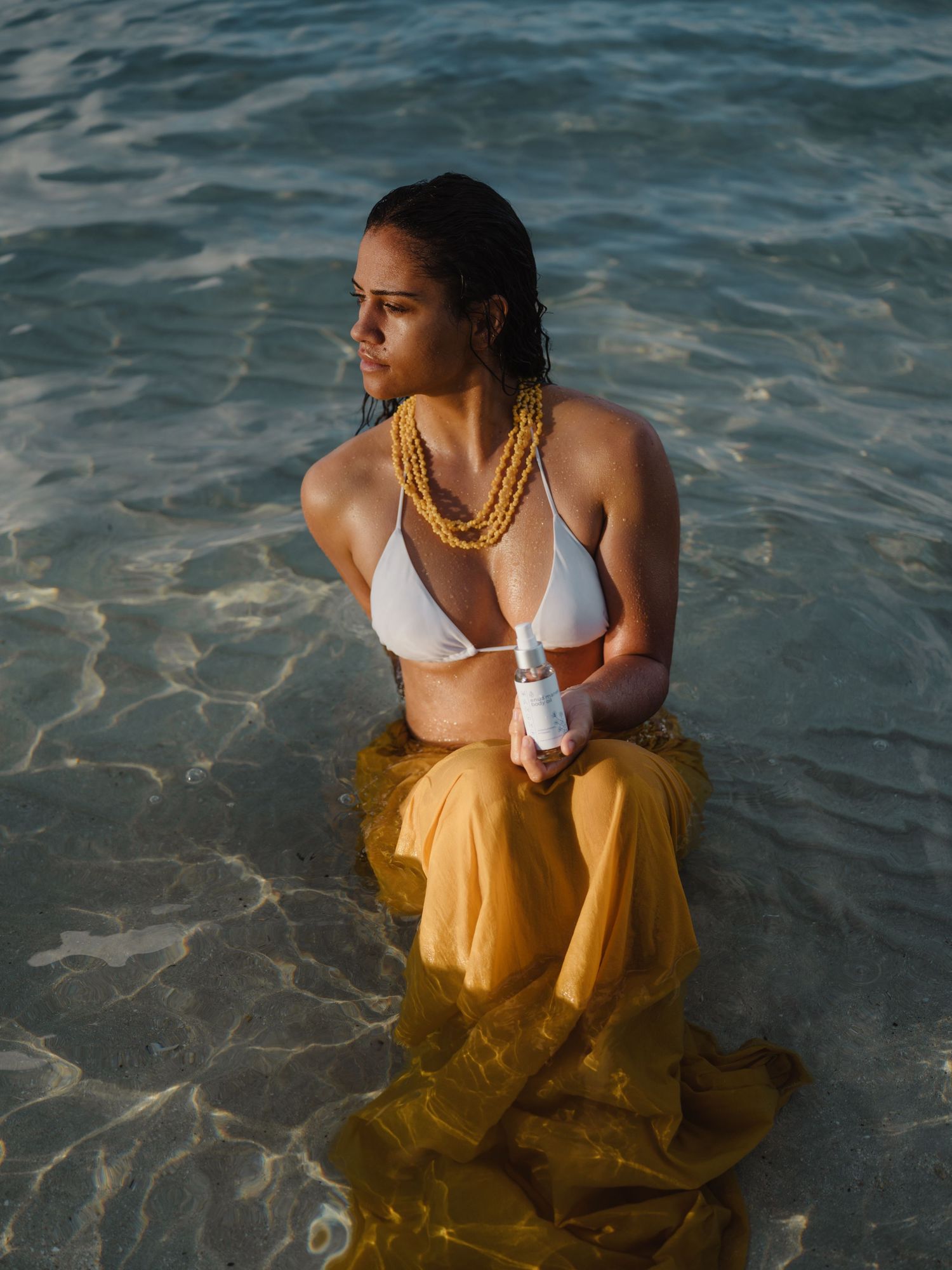 Woman in white bikini top and yellow skirt sits in shallow ocean water wearing gold layered necklaces.