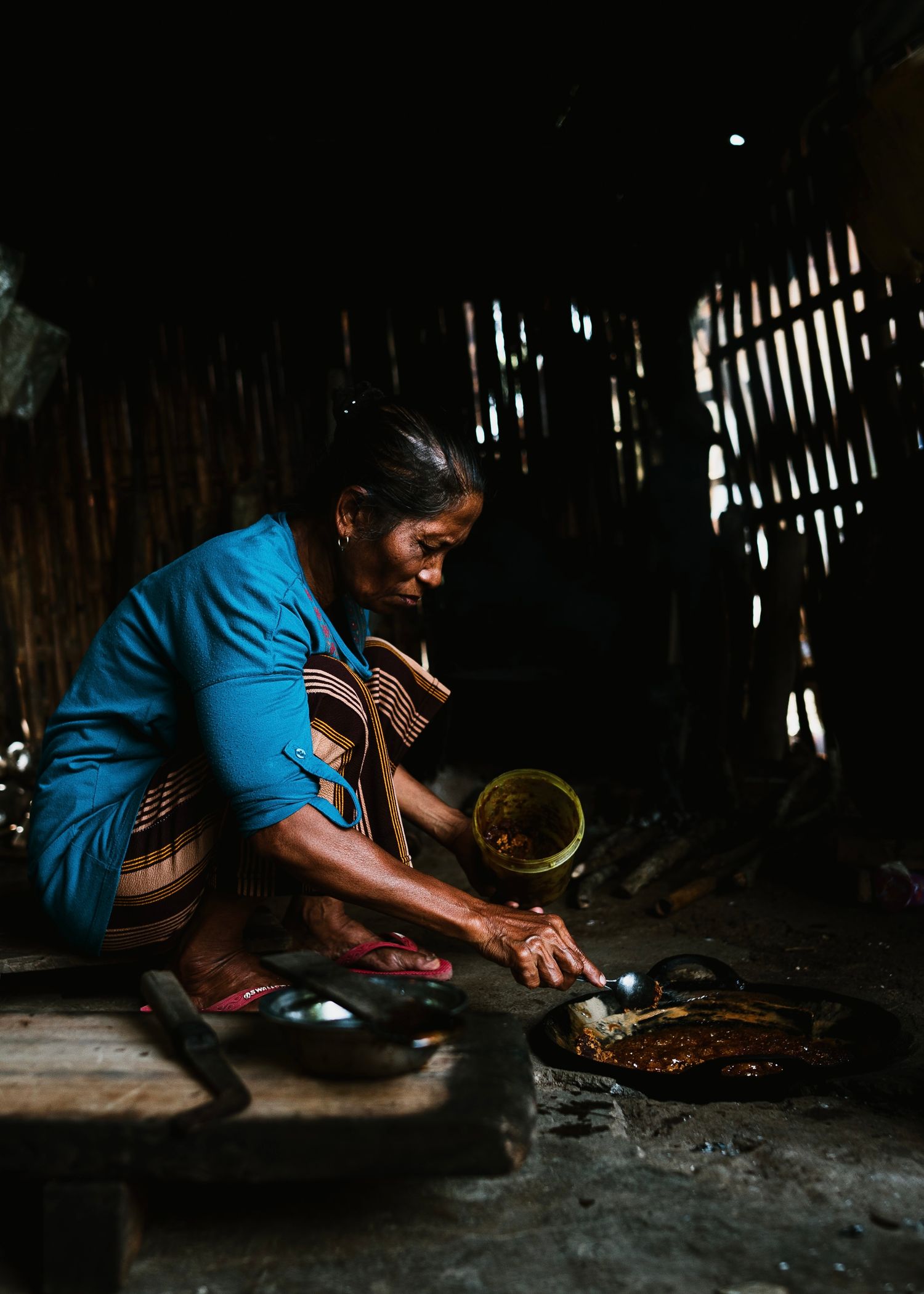 Elderly woman in blue dress cooking traditional food over open fire in rustic wooden hut kitchen.
