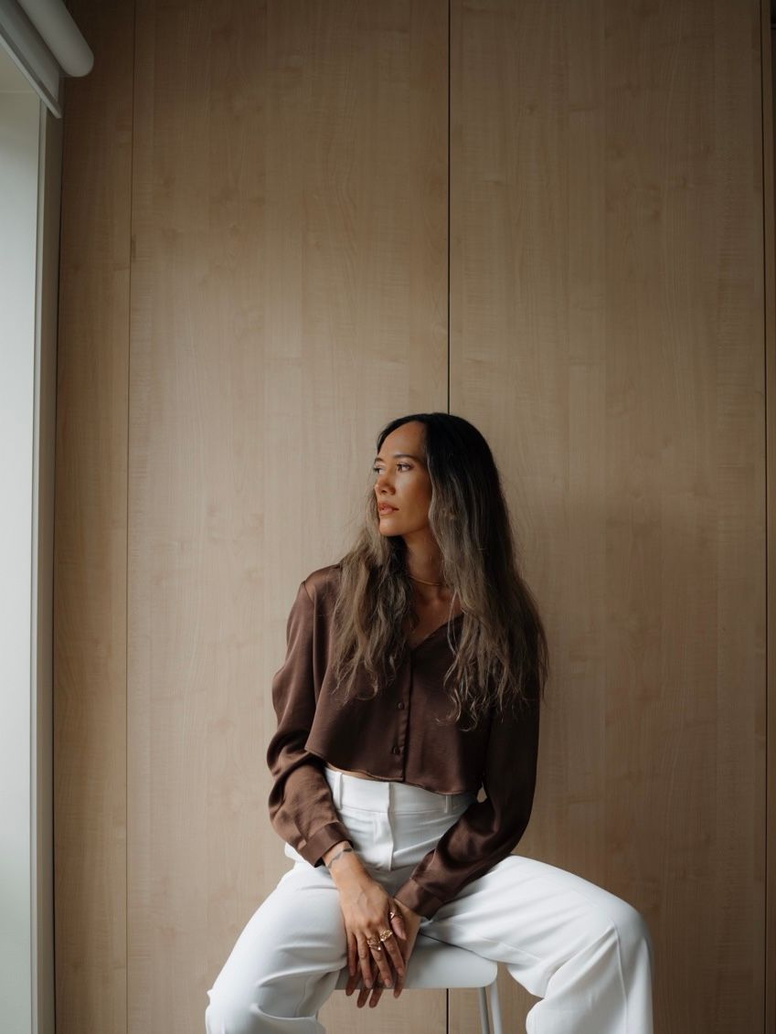 A young woman in a brown blouse and white pants sits thoughtfully against a wooden wall in soft natural light.