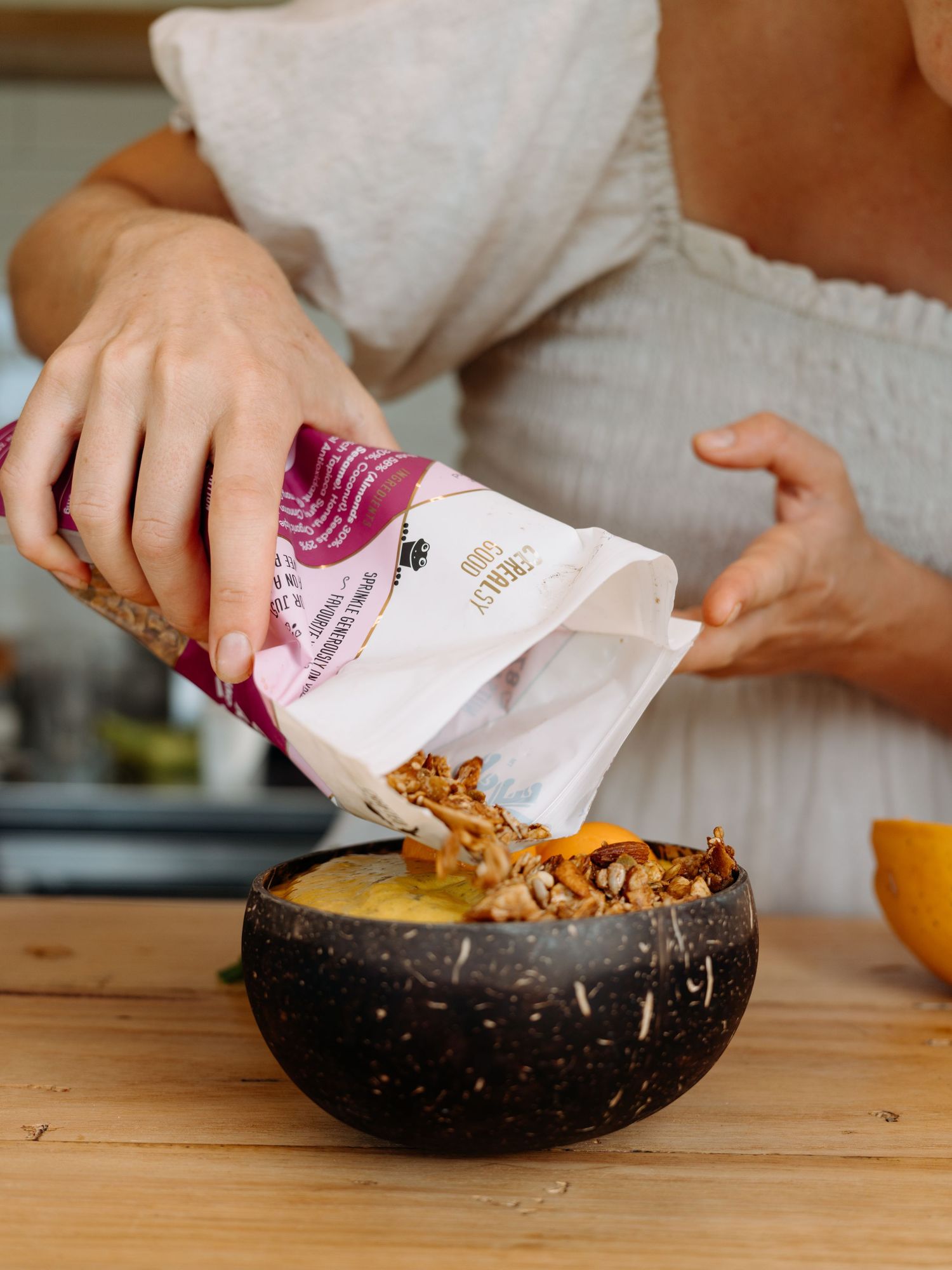 Person pouring granola from a bag into a coconut bowl filled with mango puree on a wooden table.