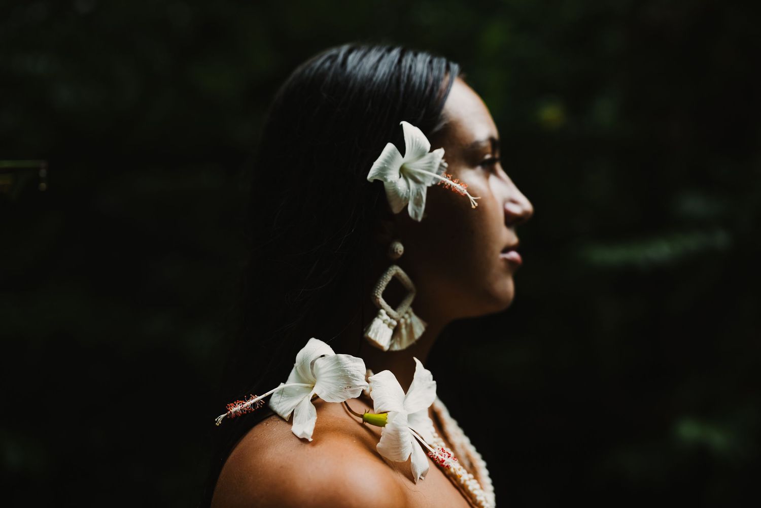 Profile of a woman wearing white floral hair accessory, matching earrings, and flower necklace against dark foliage.