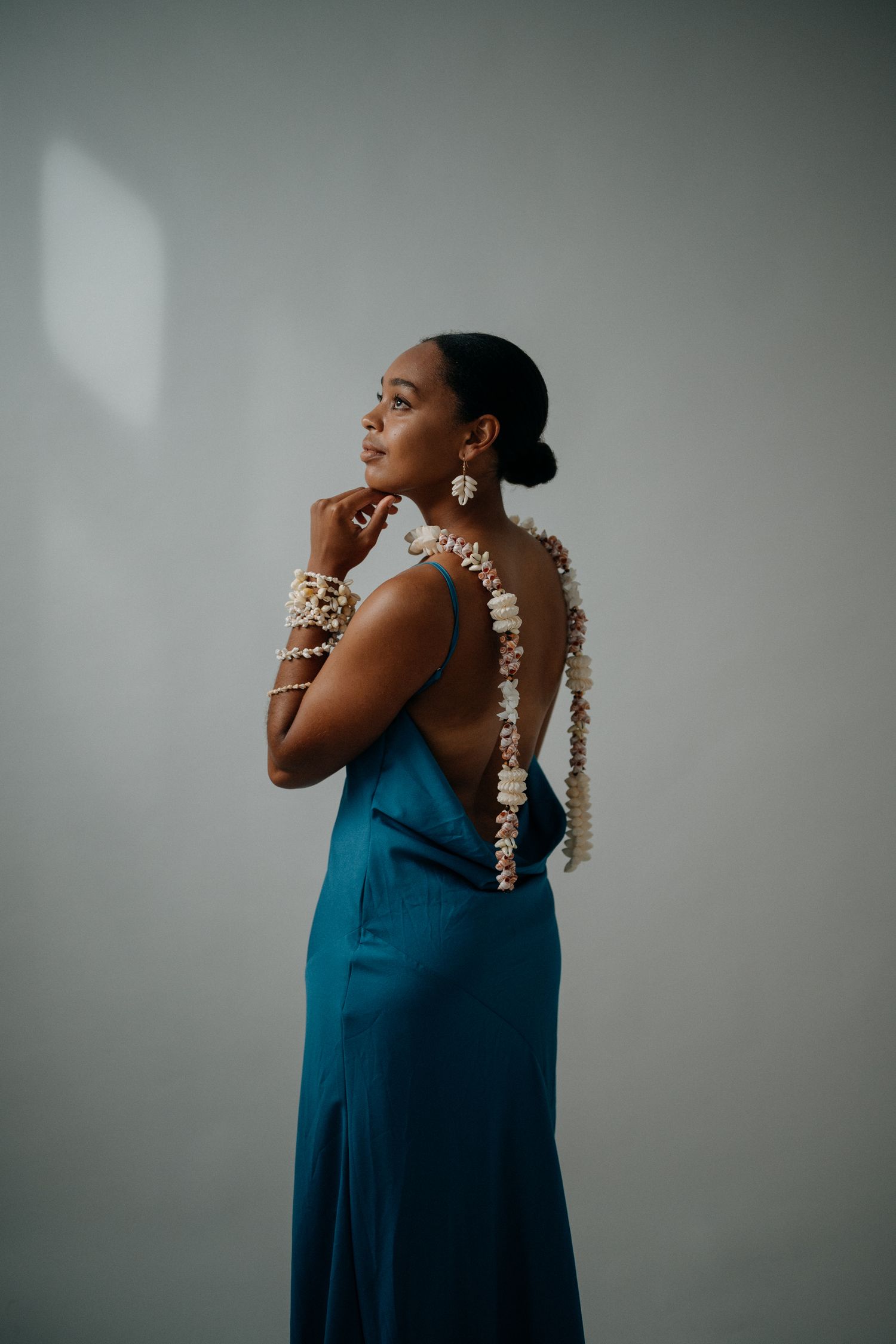 Woman in teal backless gown with floral earrings and bracelets poses gracefully against a light gray background.