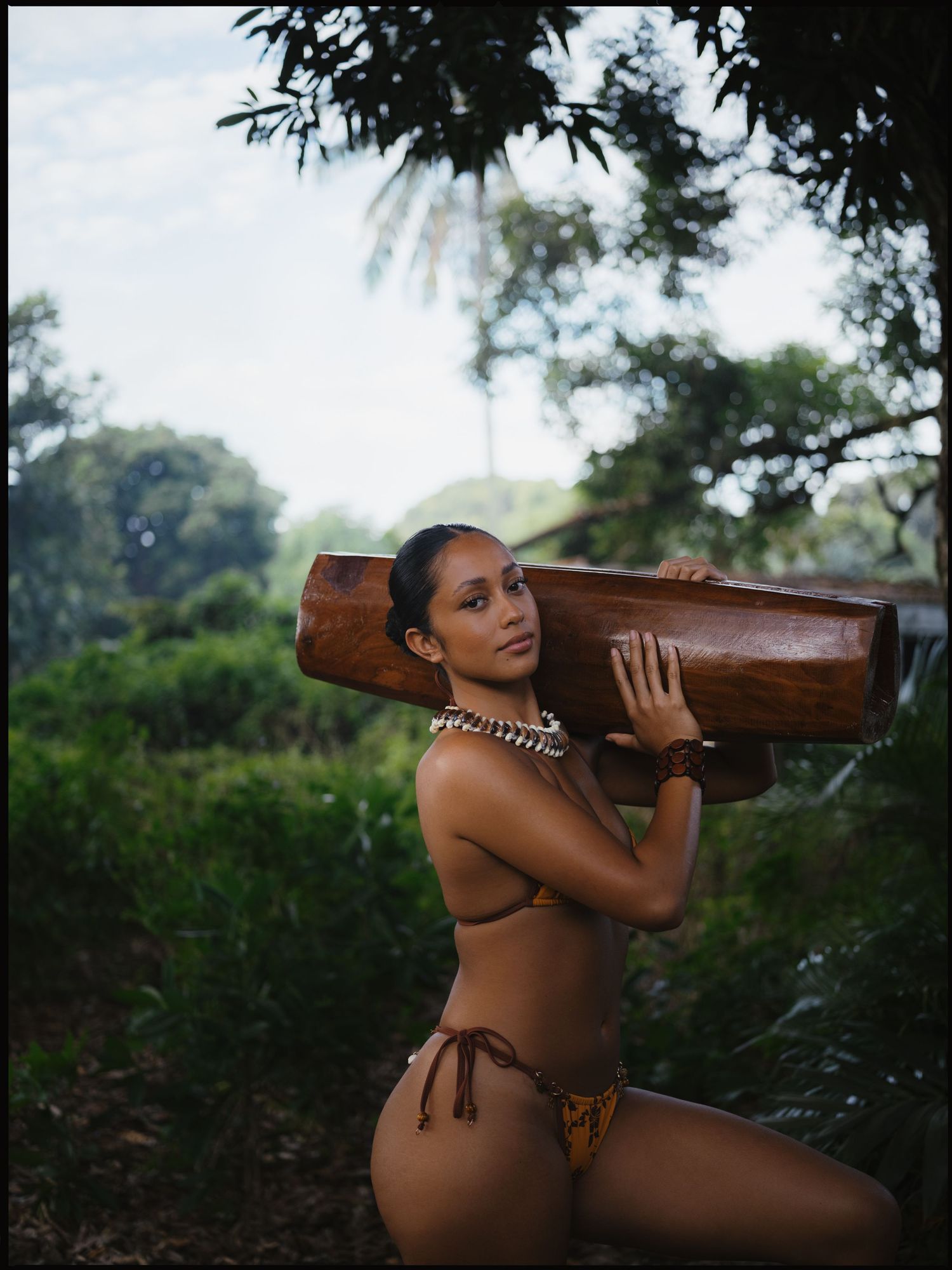 Woman in bikini carrying a wooden canoe on her shoulder in a lush tropical forest setting.
