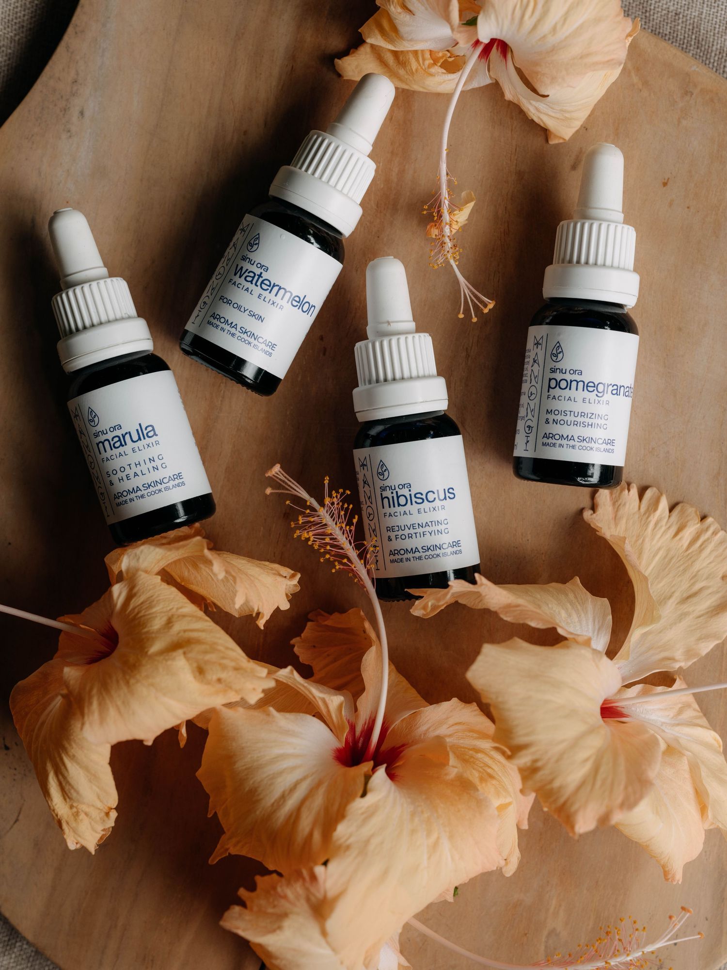 Four botanical facial oil dropper bottles with marula, hibiscus, pomegranate, and watermelon surrounded by peach hibiscus flowers.
