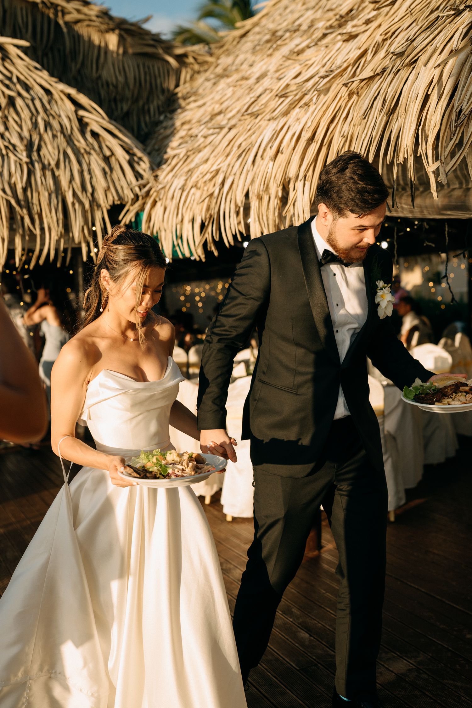 Bride in white gown and groom in black tuxedo carry plates at outdoor tropical wedding reception with thatched roof.
