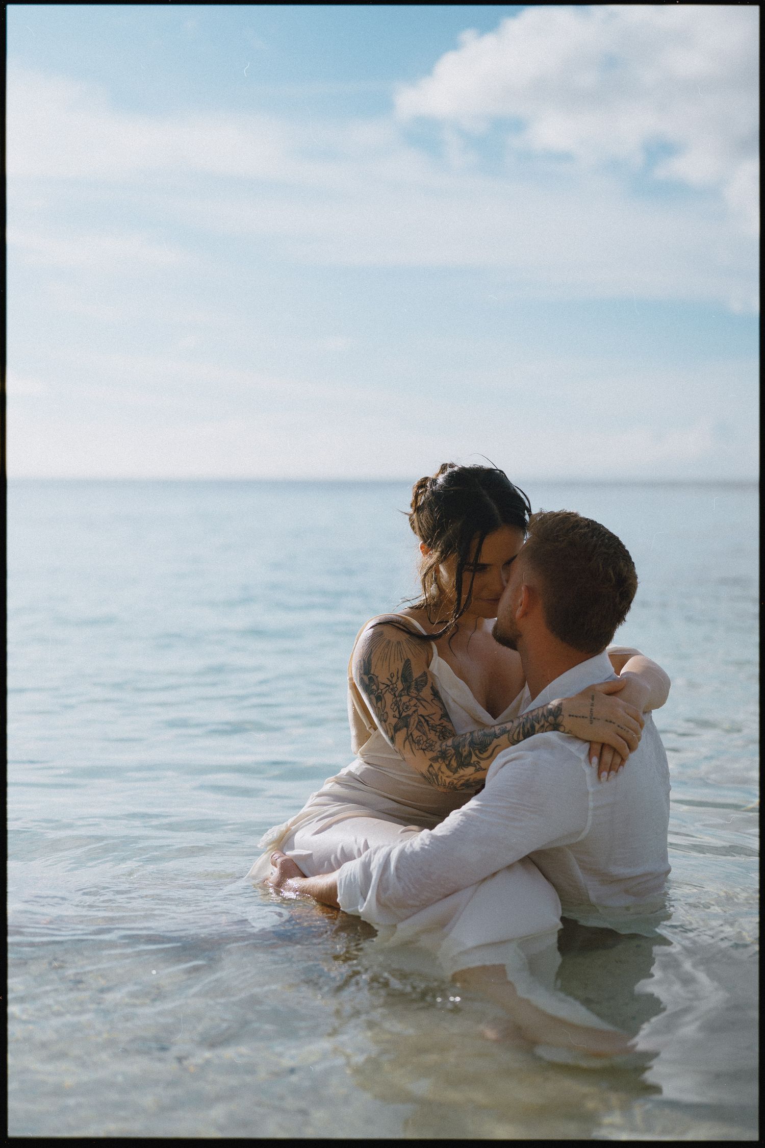 Couple embracing romantically in shallow ocean water, woman with tattoos in white dress, man in white shirt.