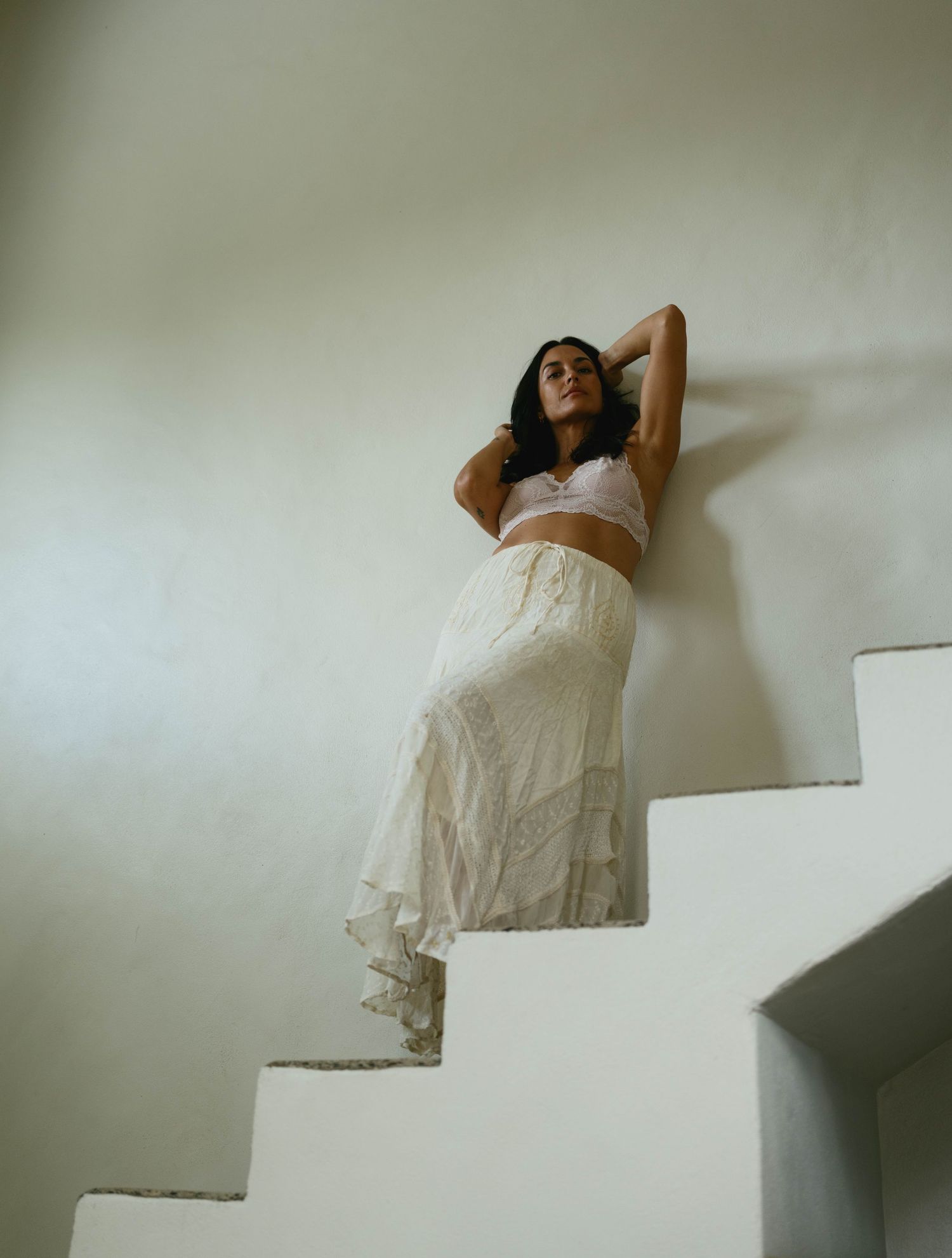 Woman in white crop top and lace maxi skirt posing gracefully on white stairs against a pale wall.