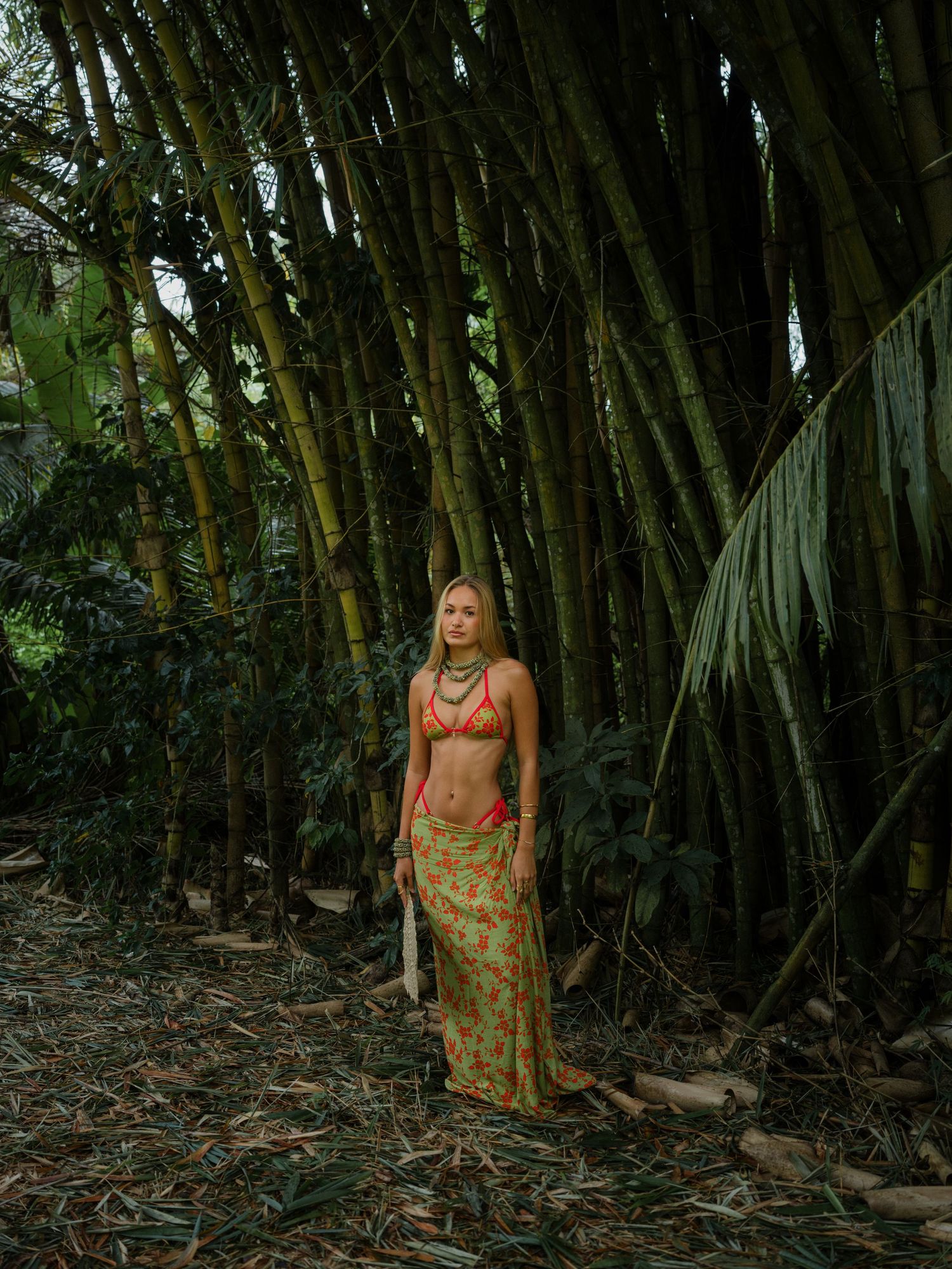 Woman in orange bikini top and floral maxi skirt standing among tall bamboo trees in a tropical forest.