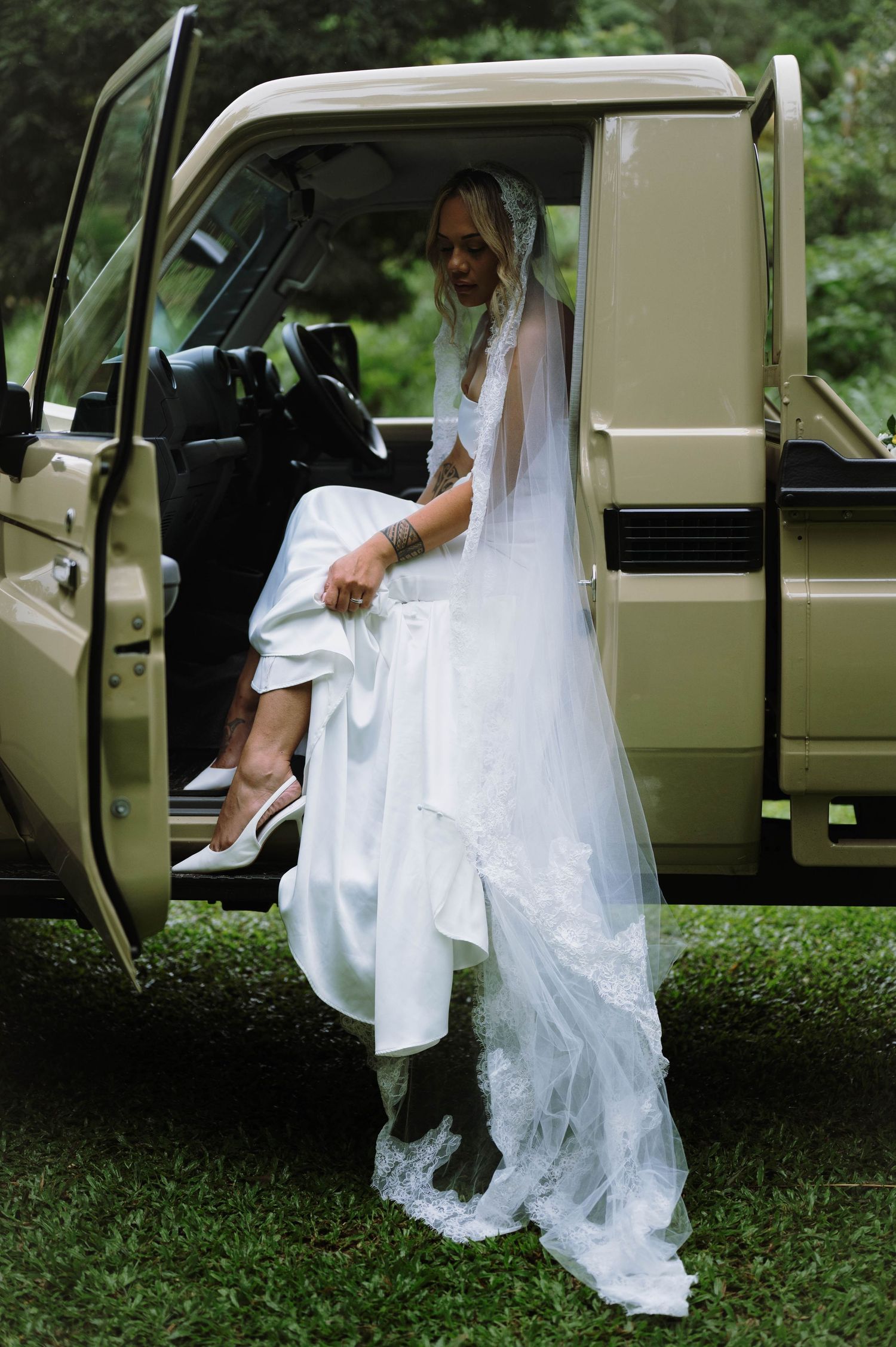 Bride in white wedding gown and veil stepping out of a vintage beige Land Rover on green grass.