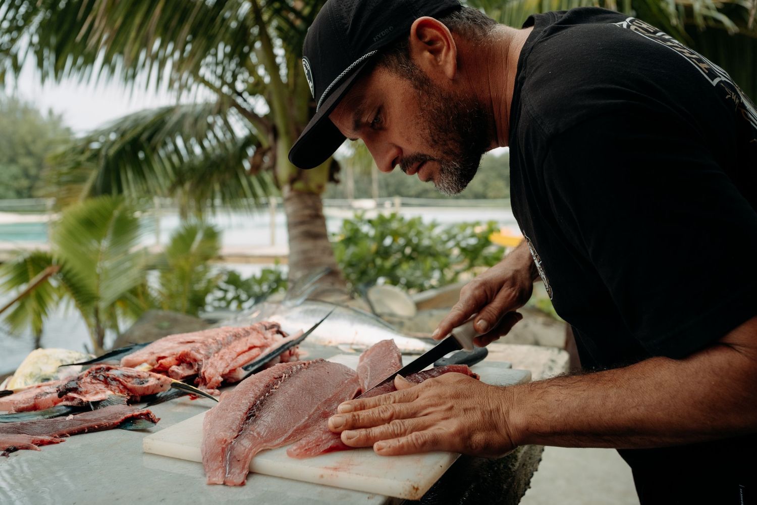 A man in a black cap fillets fresh fish on a cutting board outdoors near tropical palm trees.