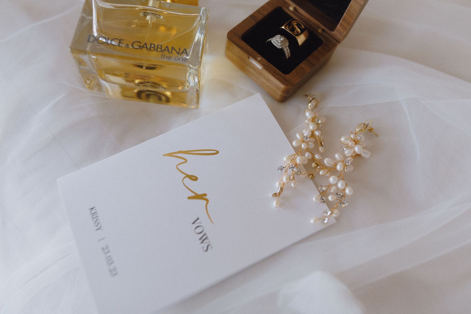 Gold wedding flat lay with Dolce Gabbana perfume, ring box, pearl earrings, and elegant card on white fabric.