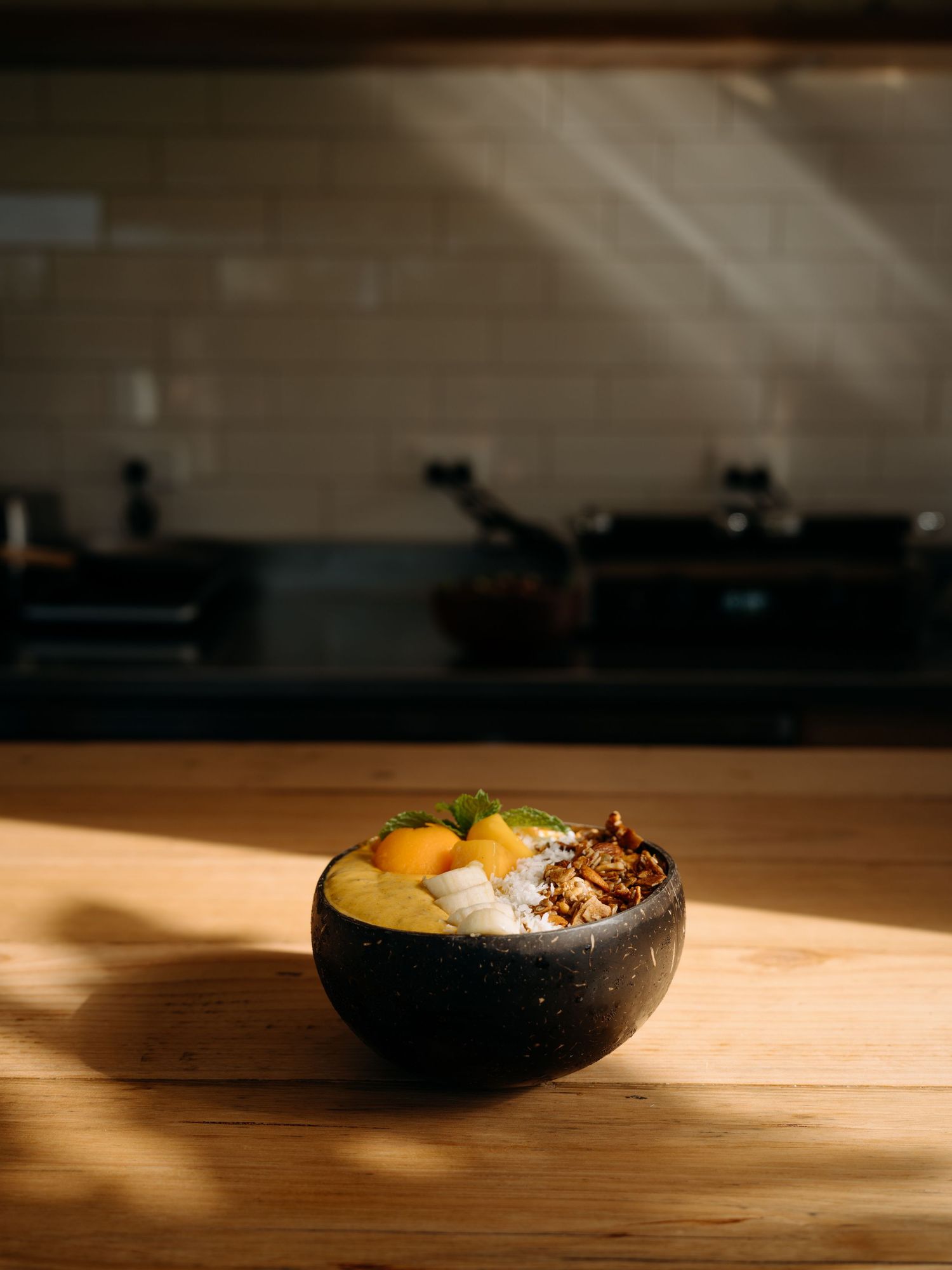 A dark coconut bowl filled with tropical fruits and granola sits on a wooden table in a sunlit kitchen.