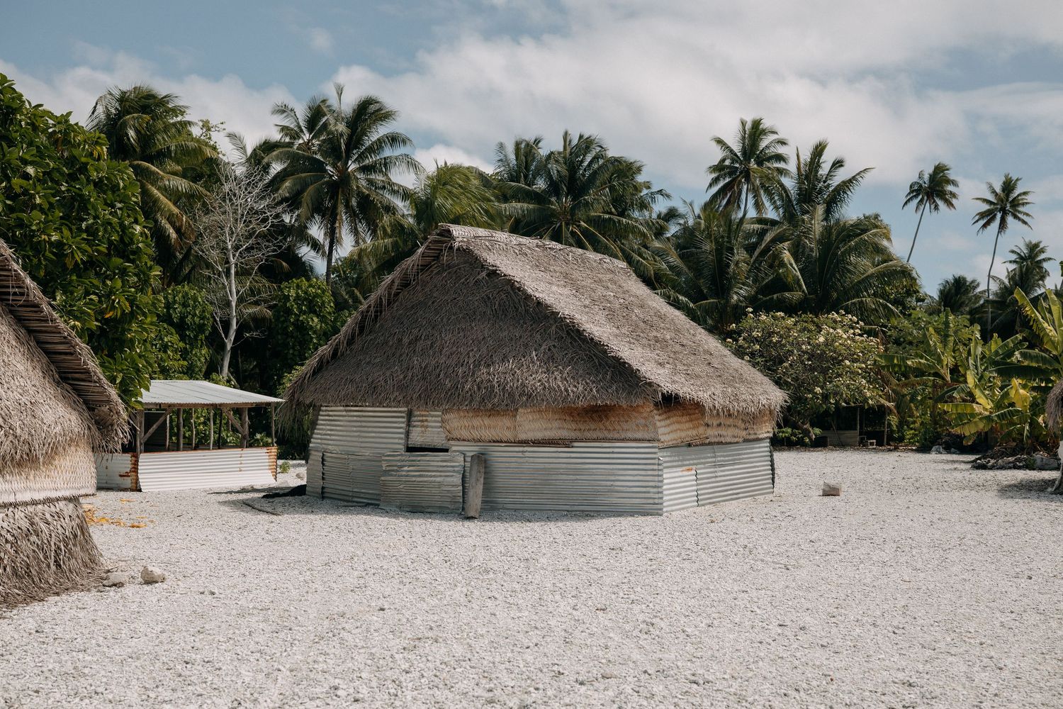 Traditional thatched roof hut on white gravel surrounded by palm trees in a tropical island setting.