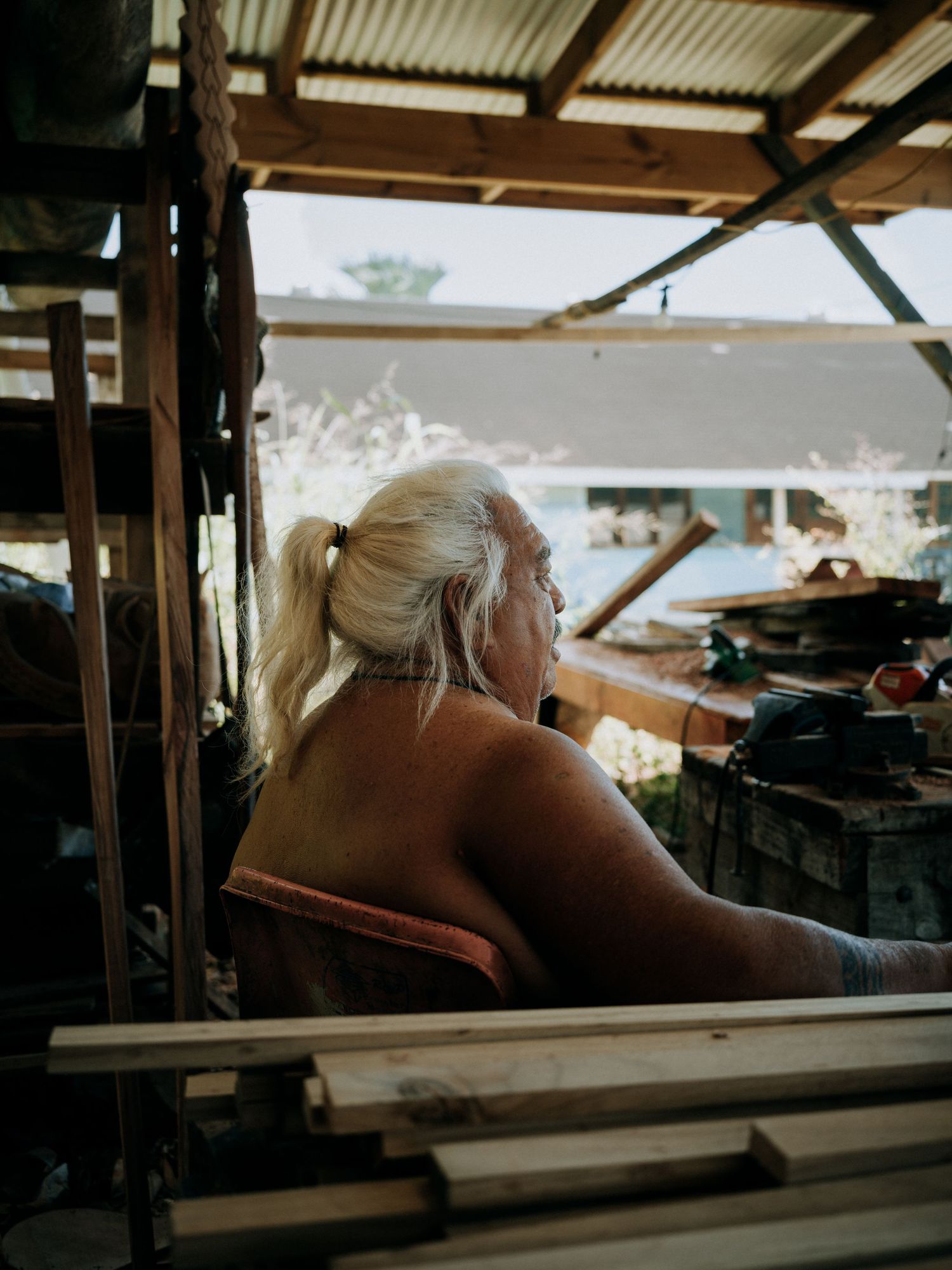 Elderly woman with white hair in ponytail sits in rustic workshop, viewed from behind, surrounded by lumber.