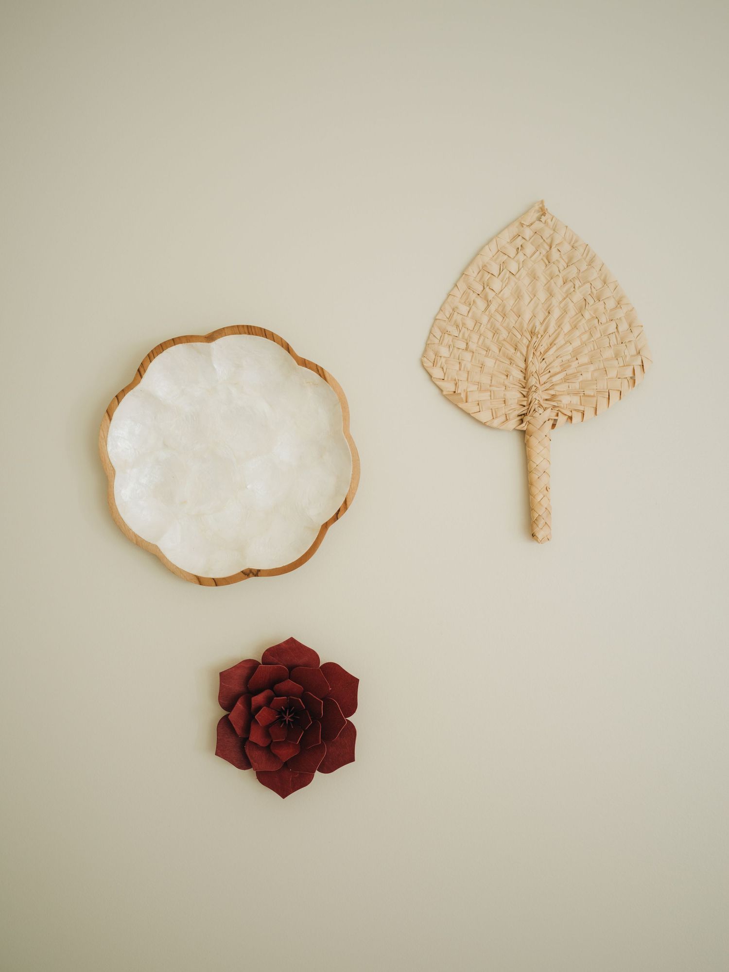 Three boho wall decor pieces: white scalloped dish, woven leaf fan, and dark red succulent on beige wall.