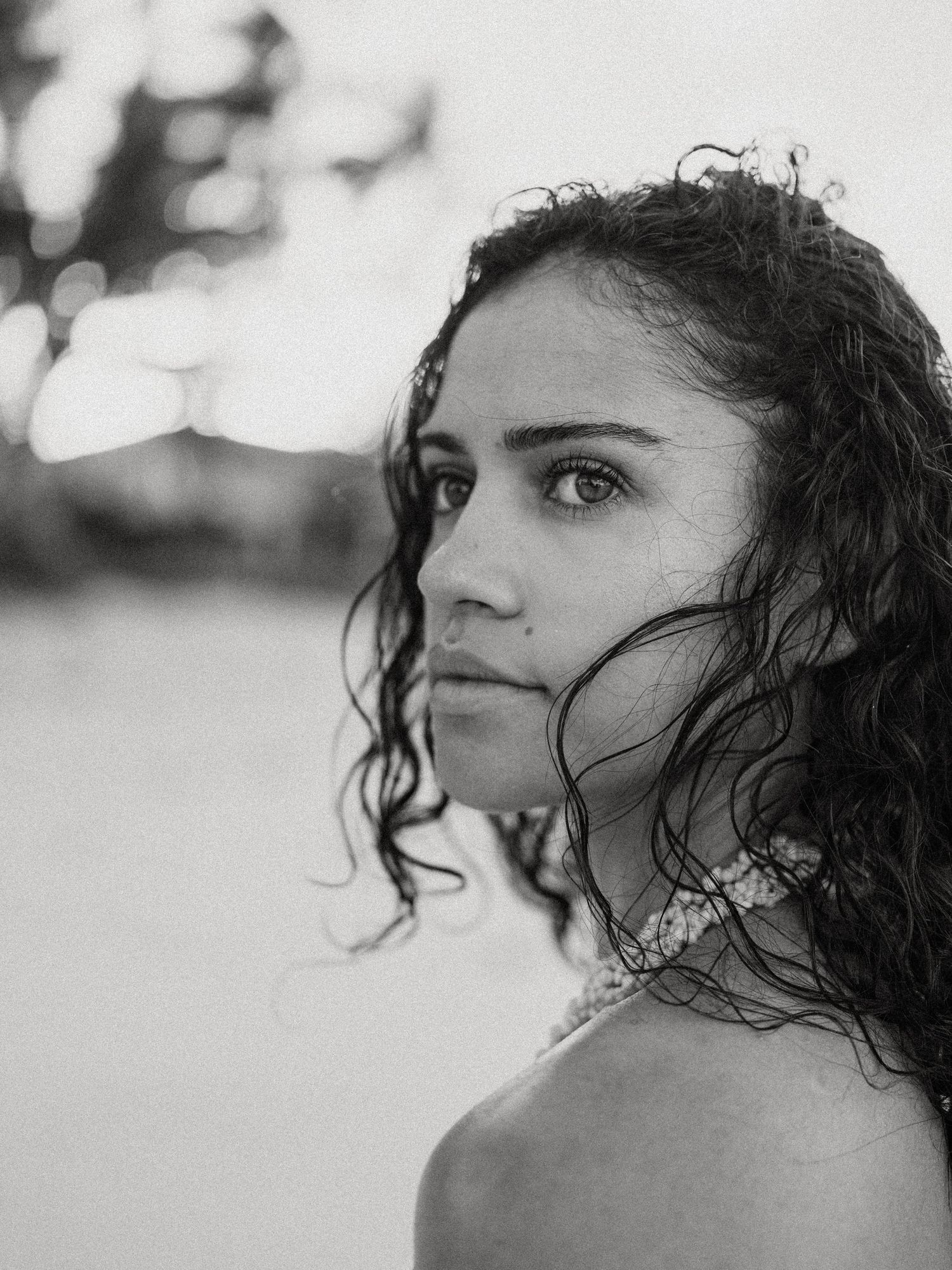 Black and white portrait of a young woman with curly hair looking over her shoulder outdoors.