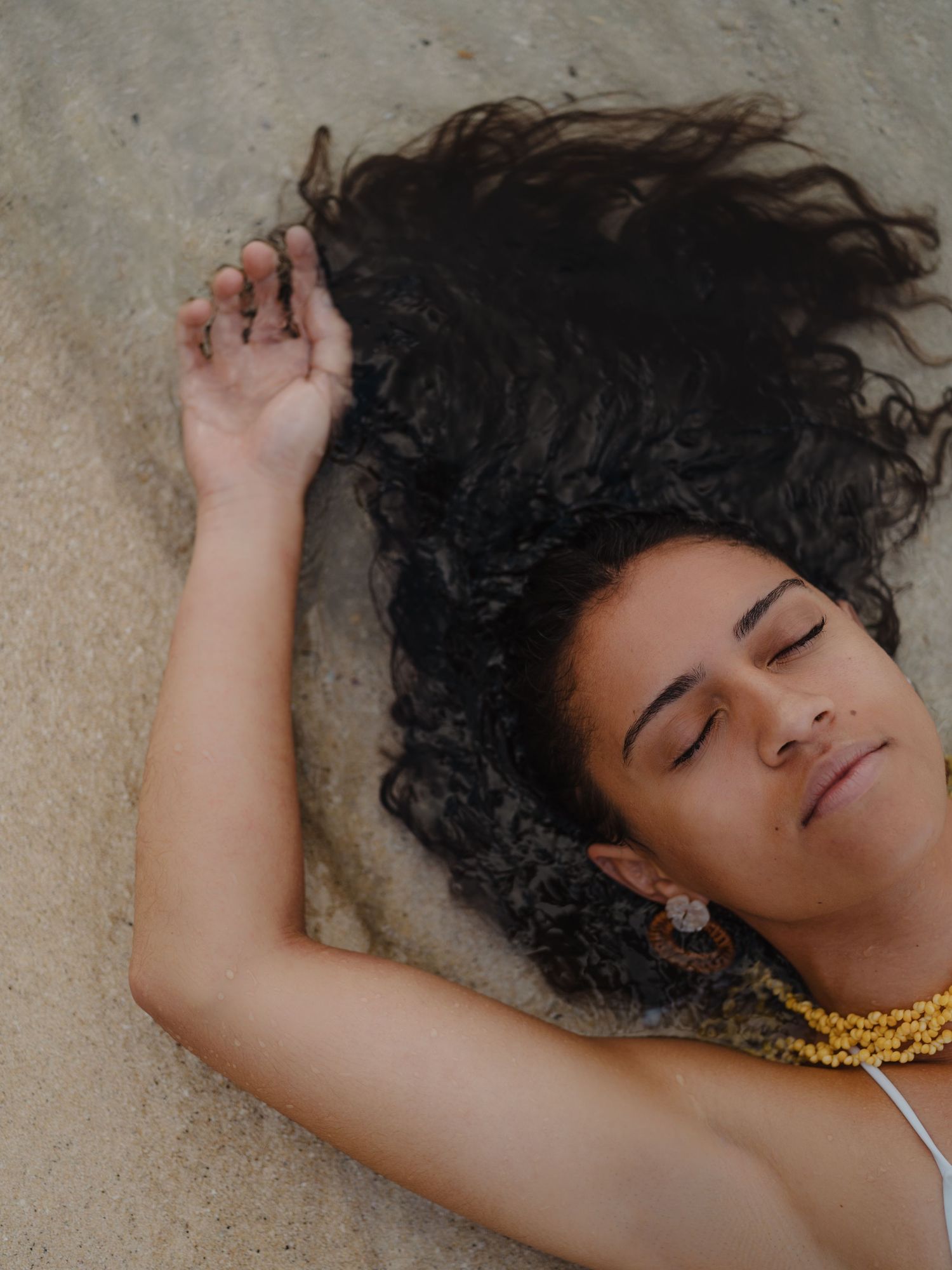 Woman with dark curly hair lying on sand beach, eyes closed, wearing yellow beaded necklace and ornate earrings.