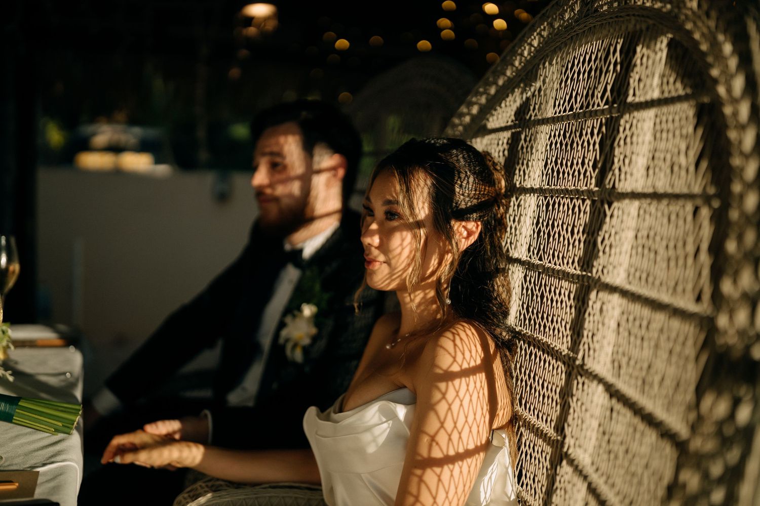 Bride and groom seated at wedding reception, bride smiling in sunlight with wicker chair shadow patterns.