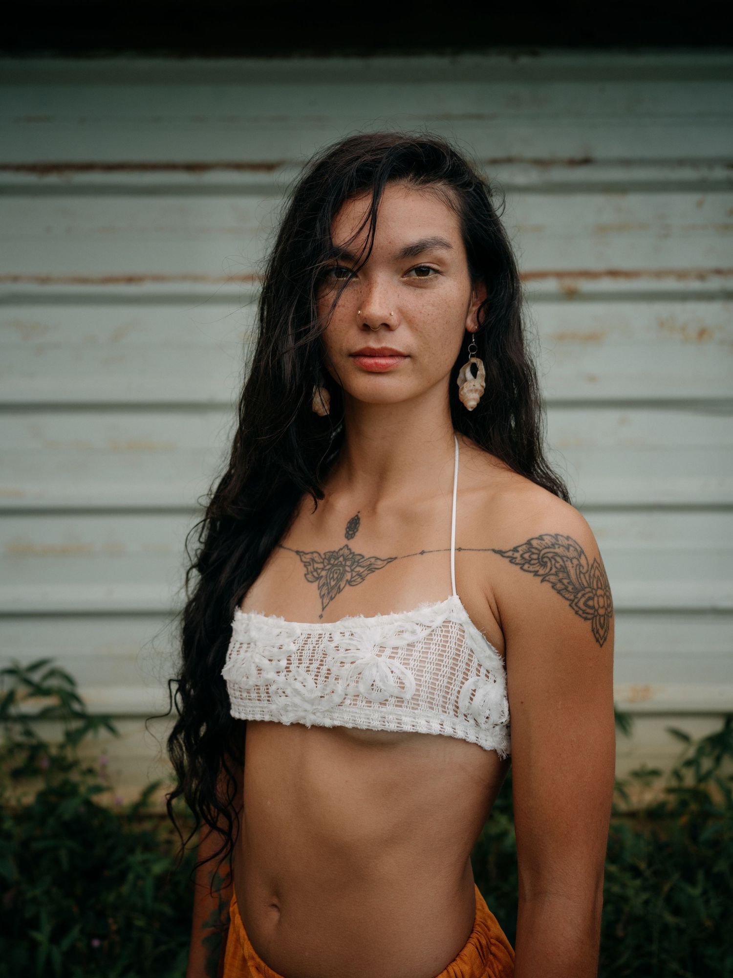 Young woman with long dark hair, tattoos, and gold earrings wearing a white crochet crop top and orange skirt outdoors.