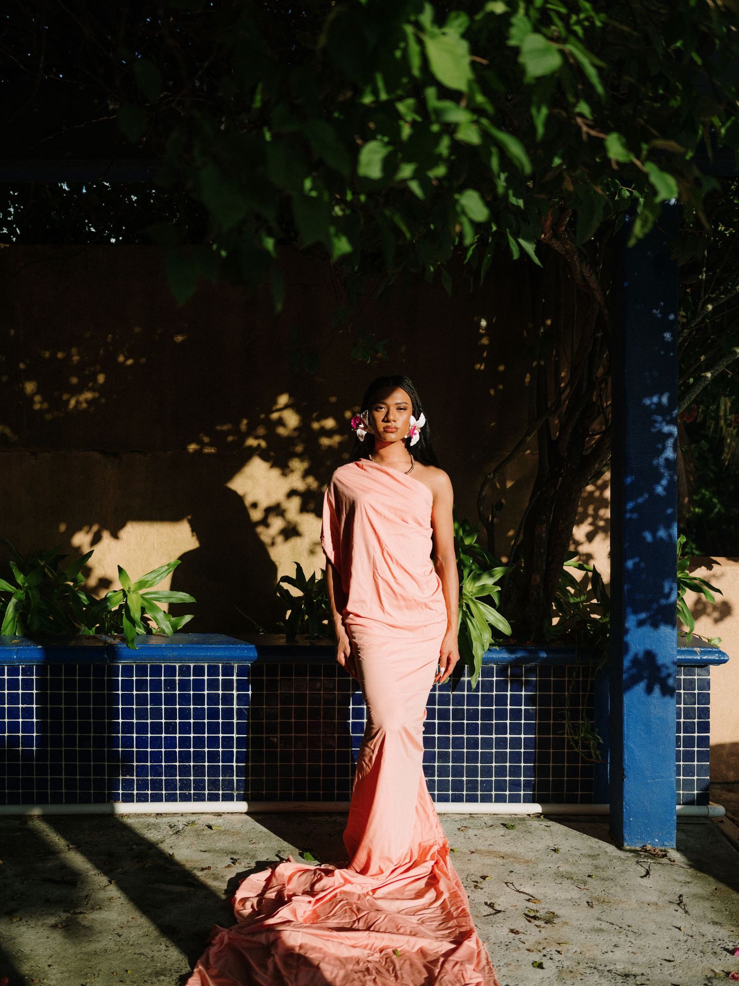Woman in elegant coral mermaid gown with floral hair accessory poses in sunlit garden with blue tile wall.