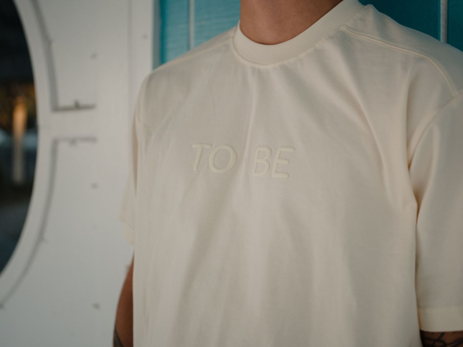 Man wearing a cream colored t-shirt with embossed TO BE text logo on the chest.
