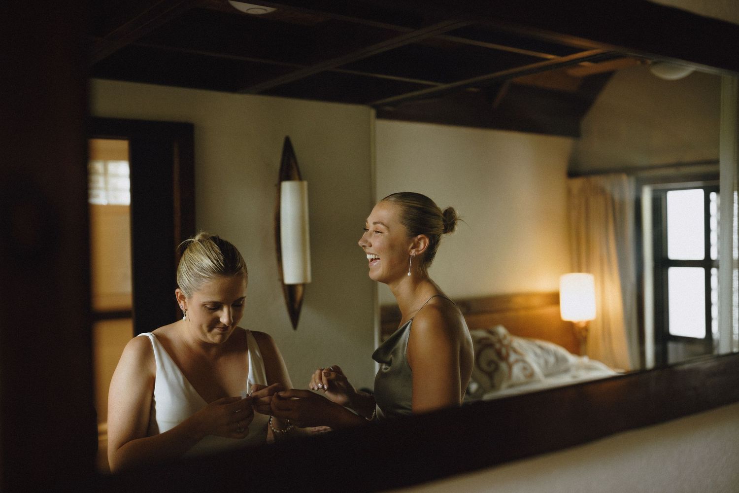 Two women laughing and sharing a joyful moment together in a warmly lit rustic bedroom.