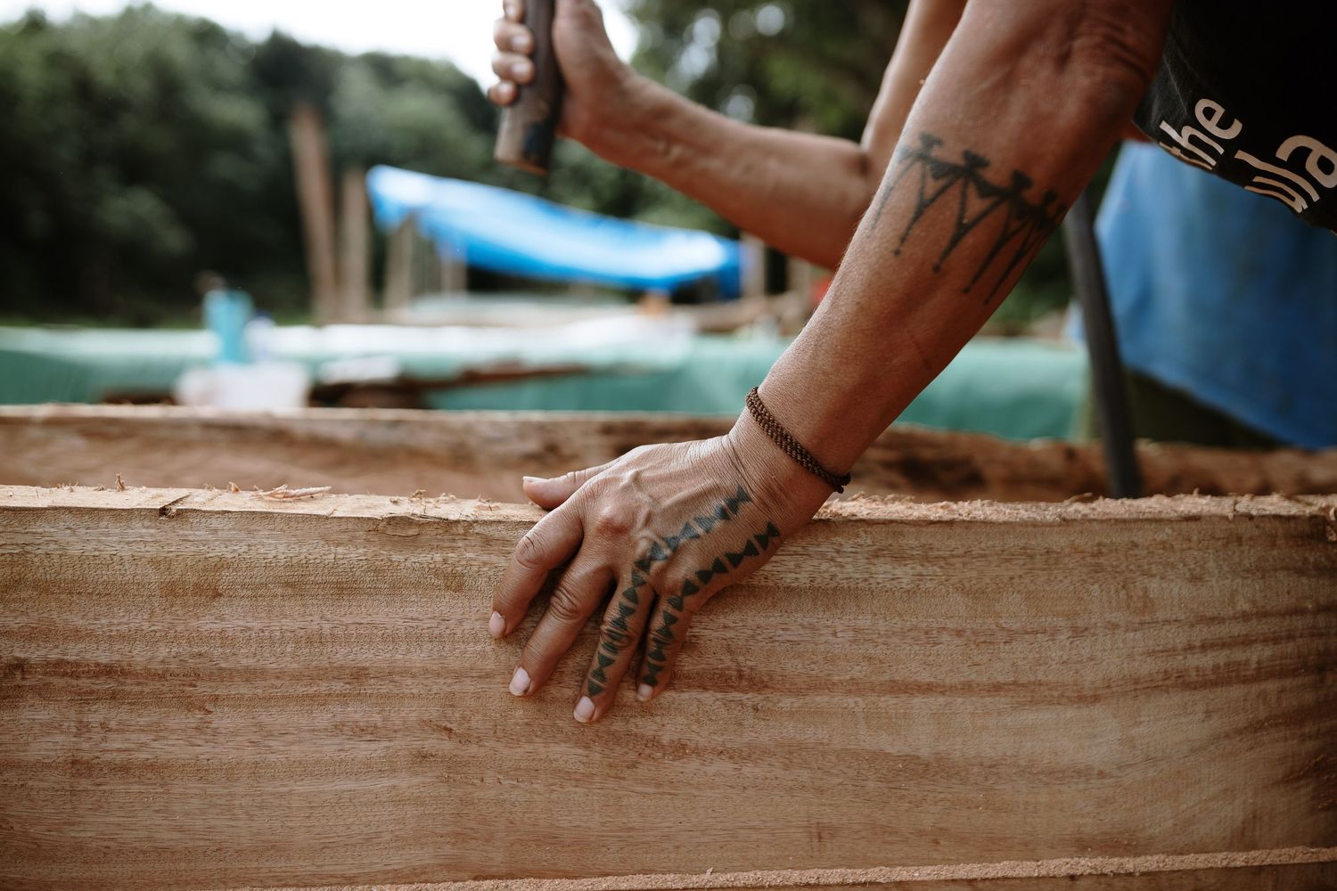 Tattooed hands of a craftsman working on wooden planks at an outdoor construction site.