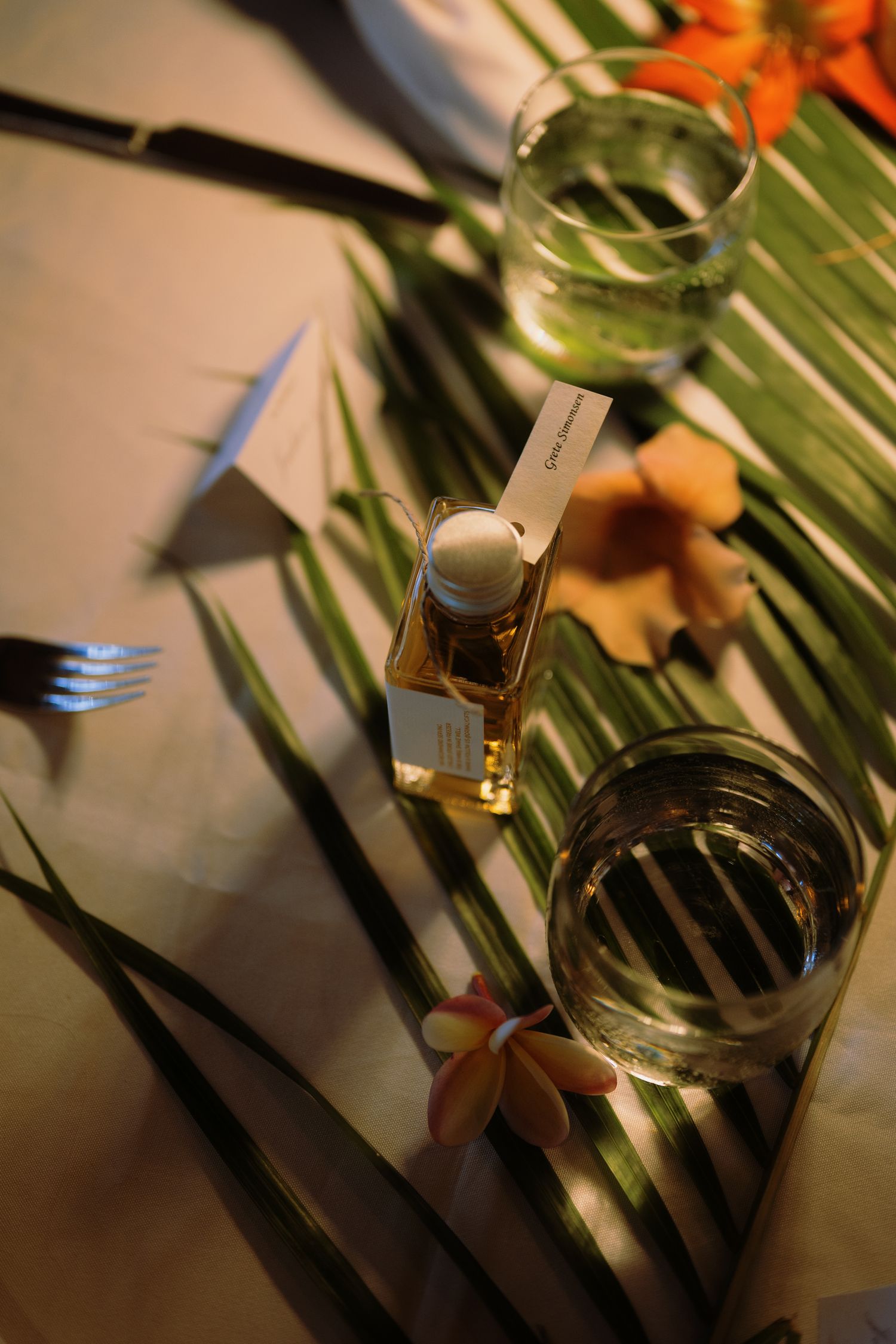 Tropical table setting with palm leaves, small perfume bottle, fork, glasses of water, and orange flowers.
