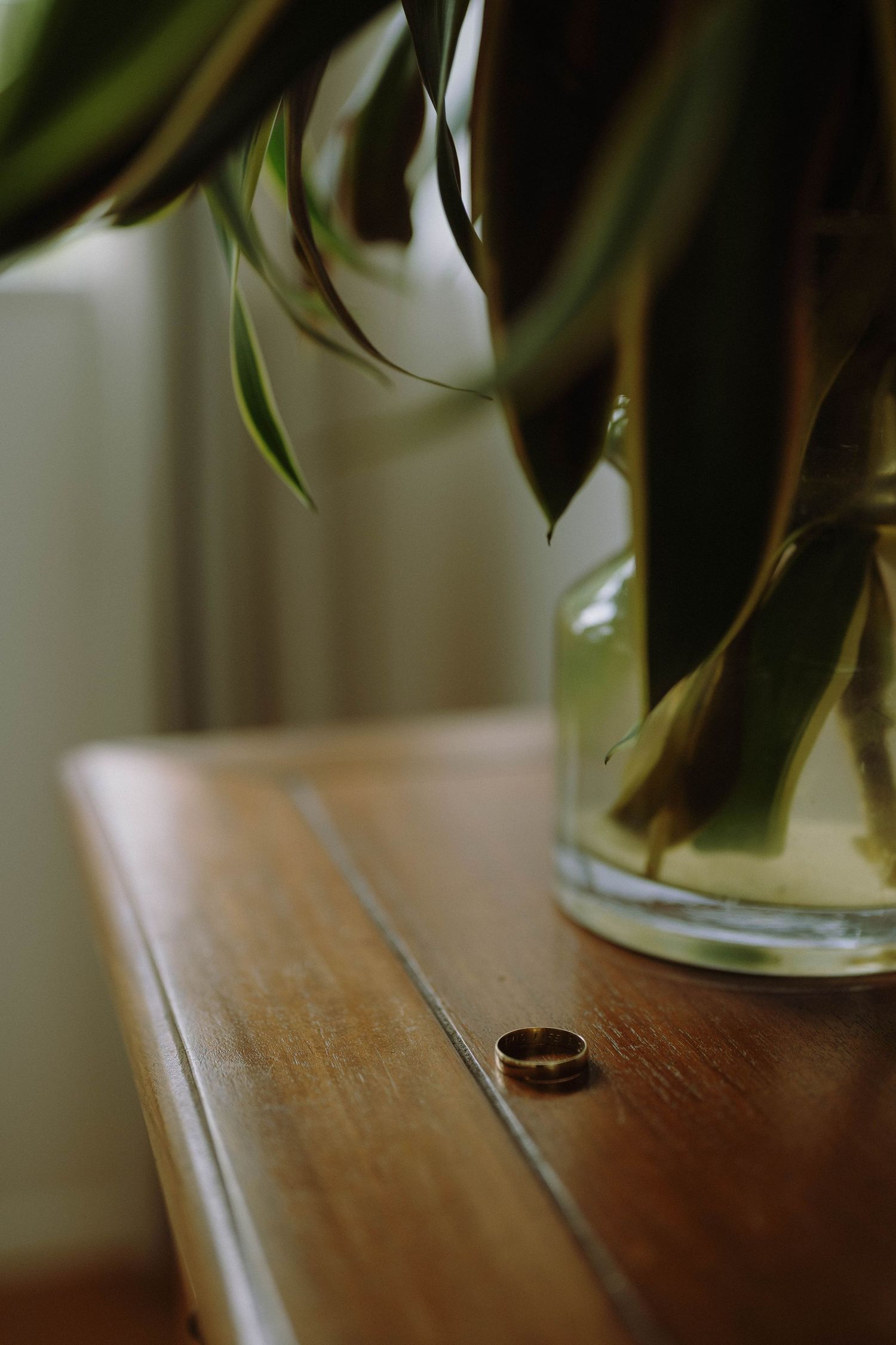 A gold wedding ring rests on a wooden dresser beside a glass vase with green tropical leaves.