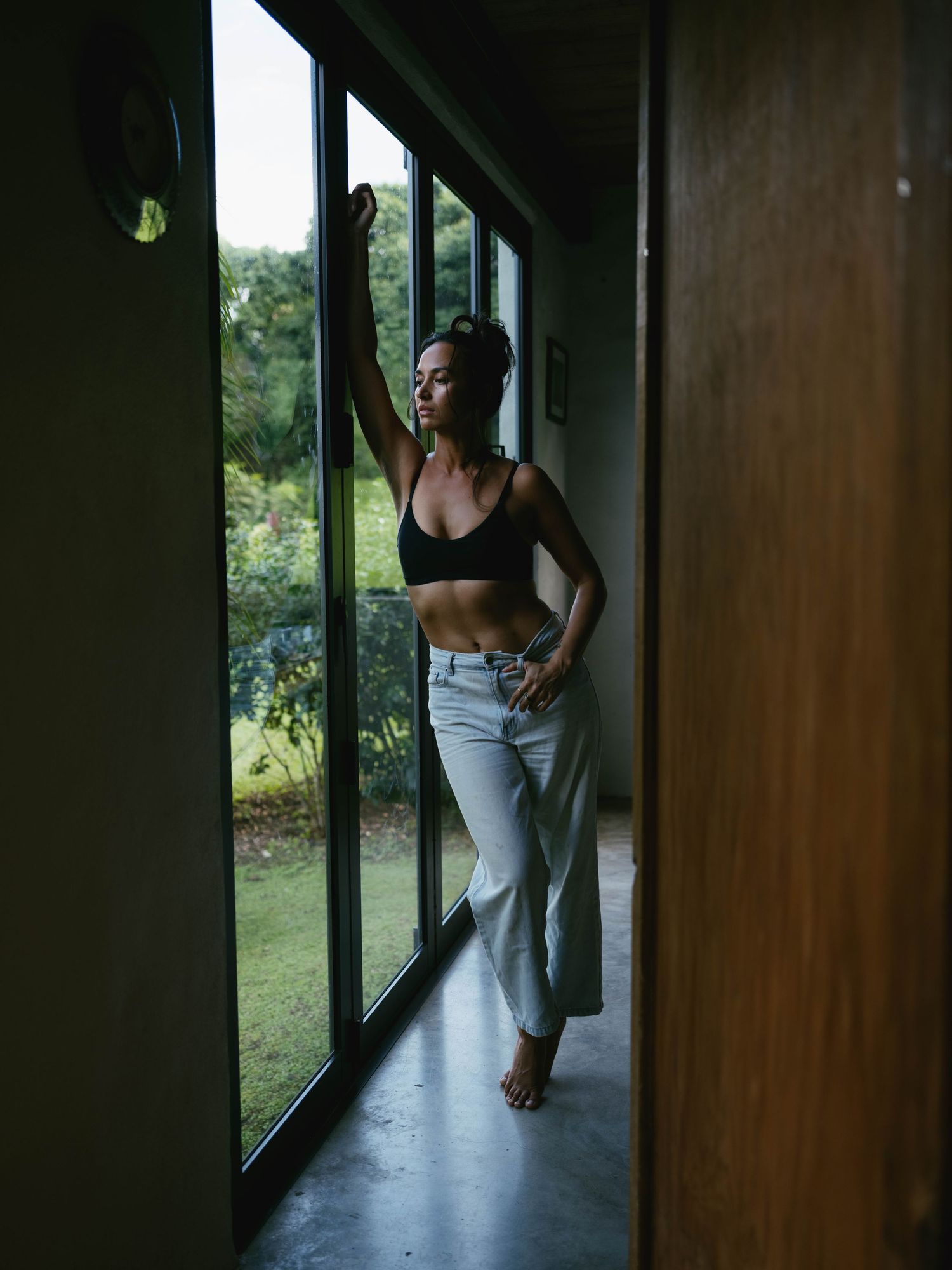 Woman in black crop top and light jeans standing barefoot by large glass doors with garden view.