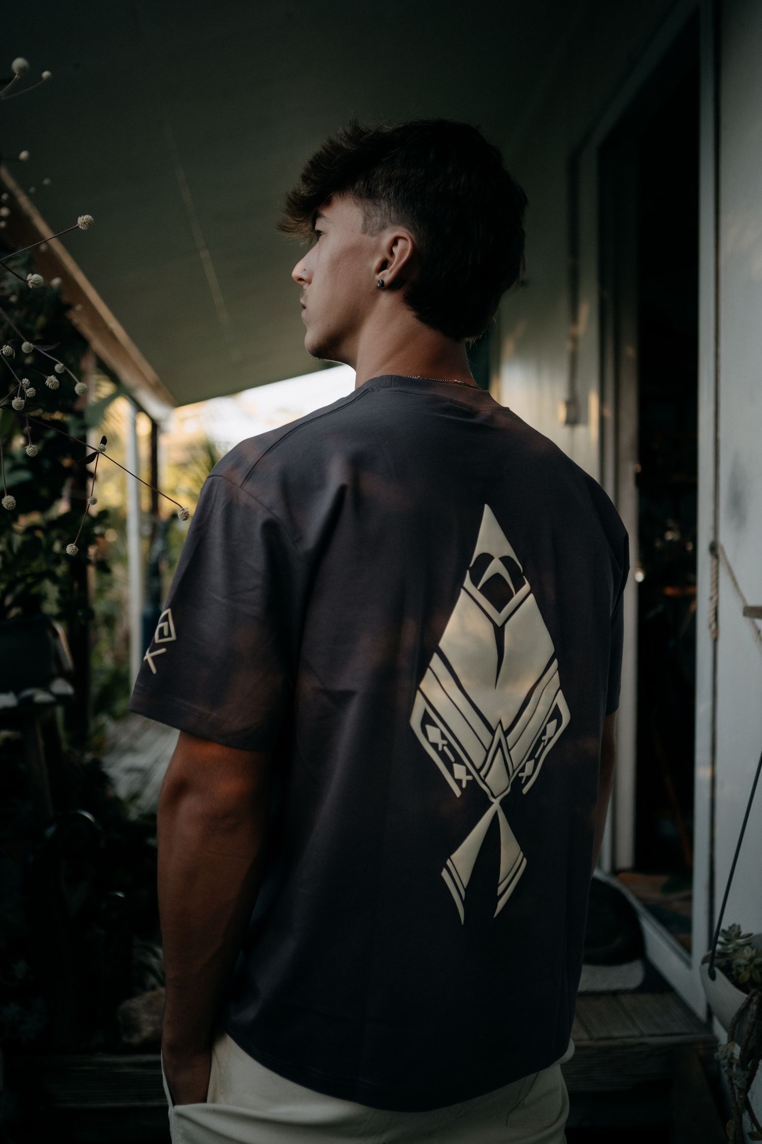 Young man in dark graphic tee with geometric back print stands on balcony surrounded by plants.