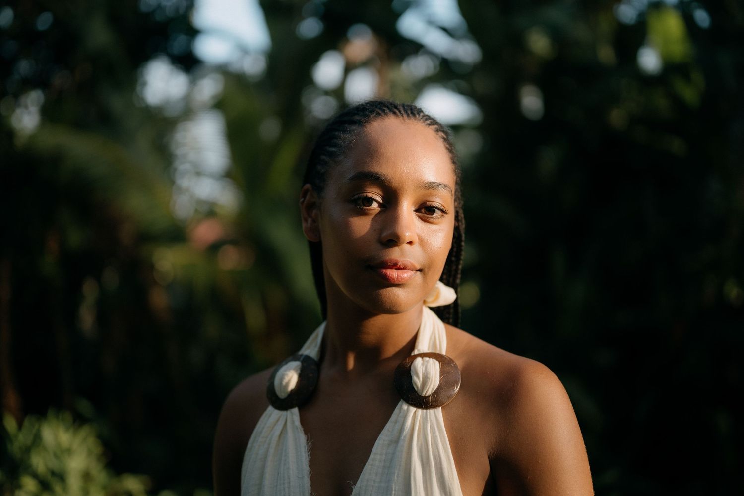 Young Black woman in white halter top wearing large hoop earrings, posing outdoors with green foliage background.