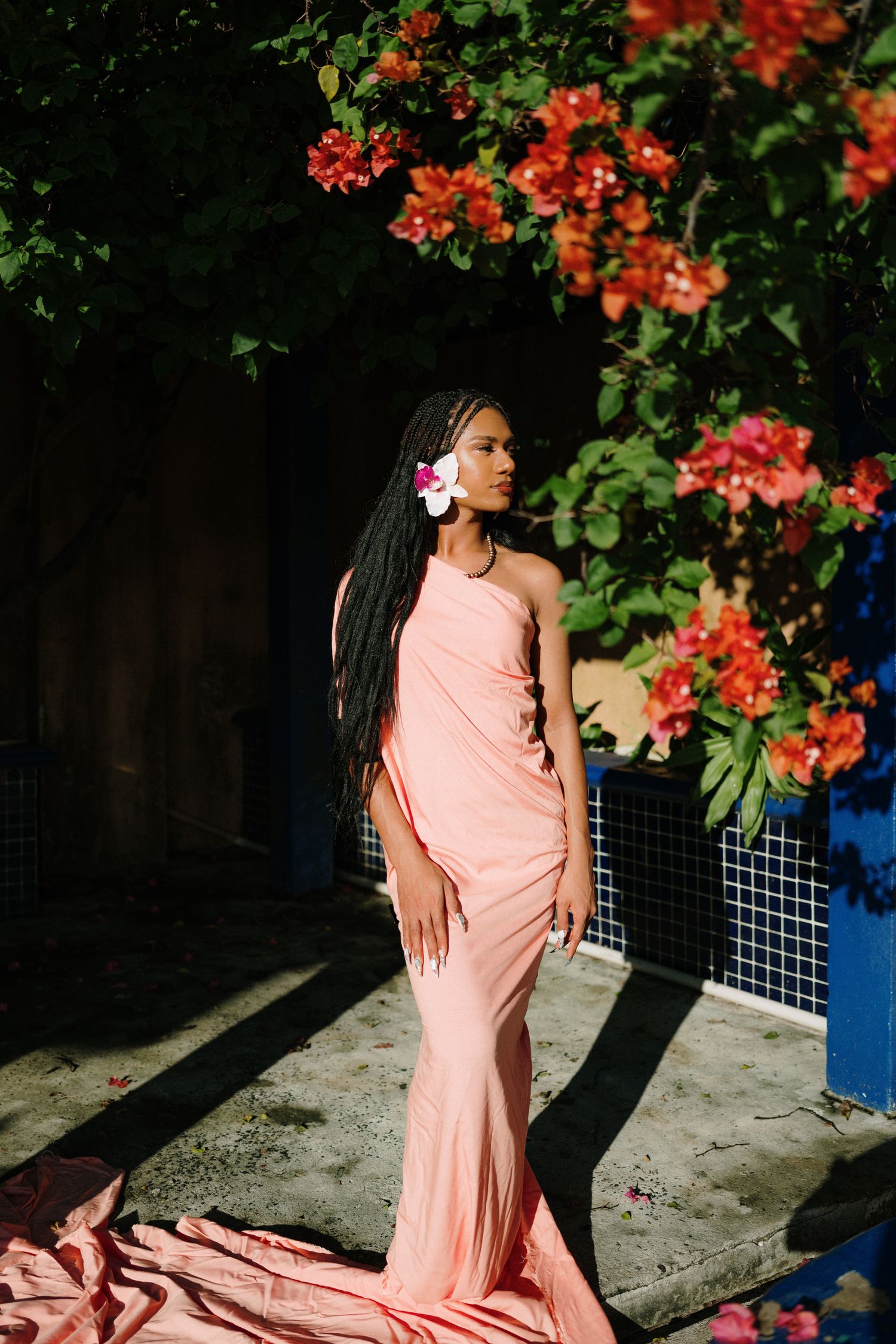 A woman in a peach gown with long braids and a flower in her hair poses near vibrant red bougainvillea blooms.