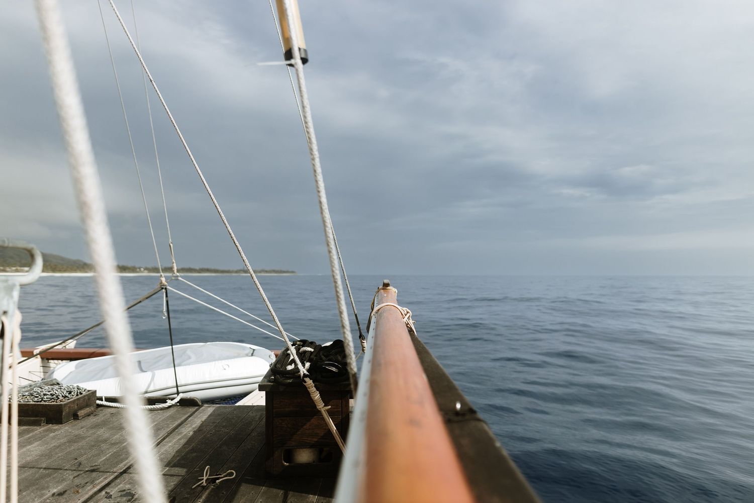 View from the bow of a sailboat on calm ocean waters under overcast skies with a small dinghy visible on deck.