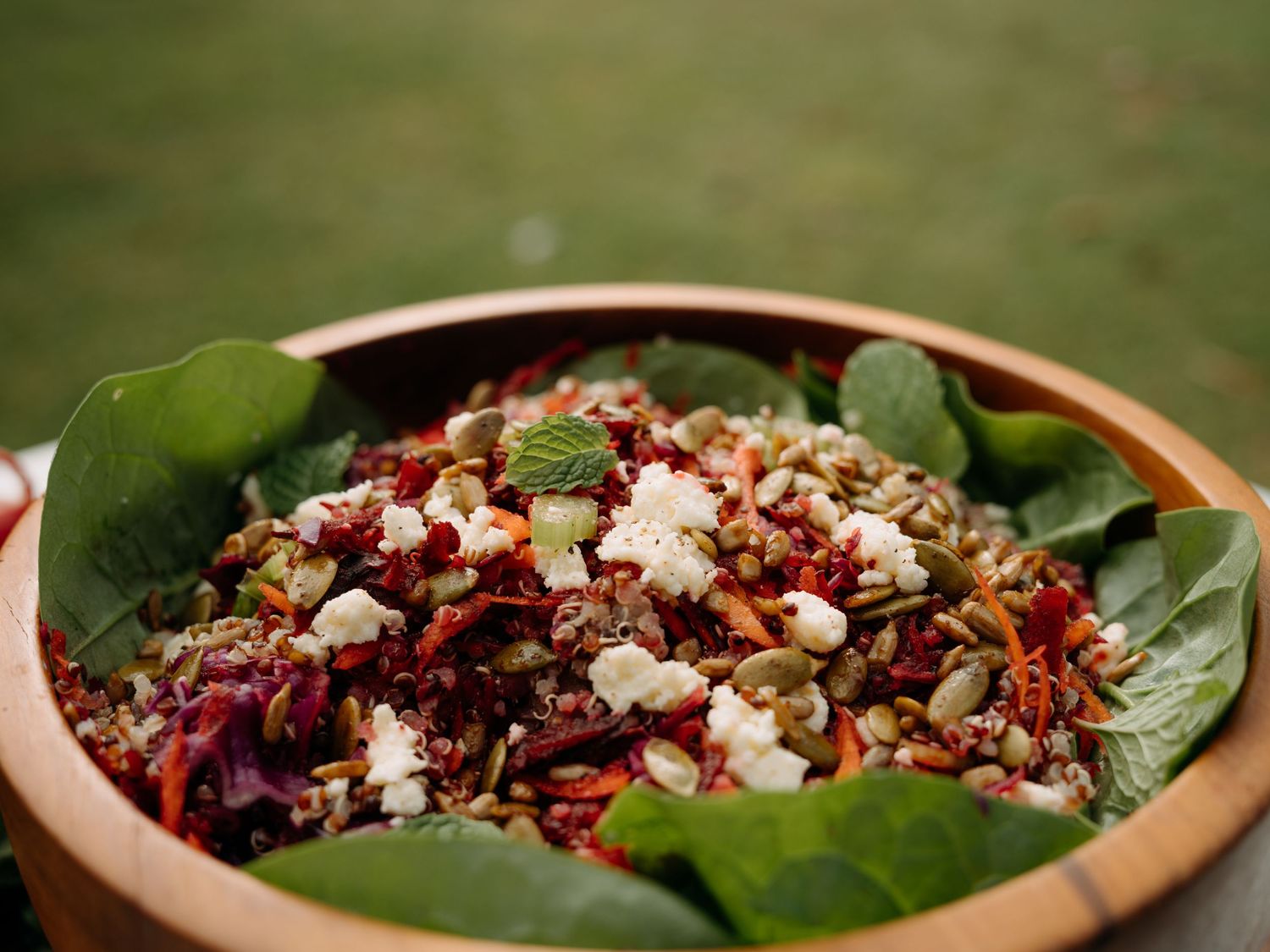Wooden bowl filled with colorful quinoa salad, fresh greens, feta cheese, seeds, and pomegranate seeds outdoors.