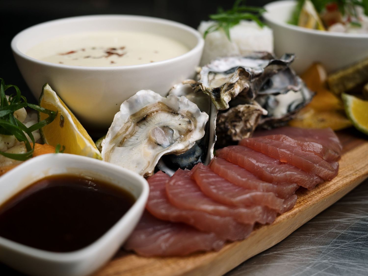 Fresh seafood platter with oysters, tuna sashimi, dipping sauces, and creamy chowder on a wooden board.