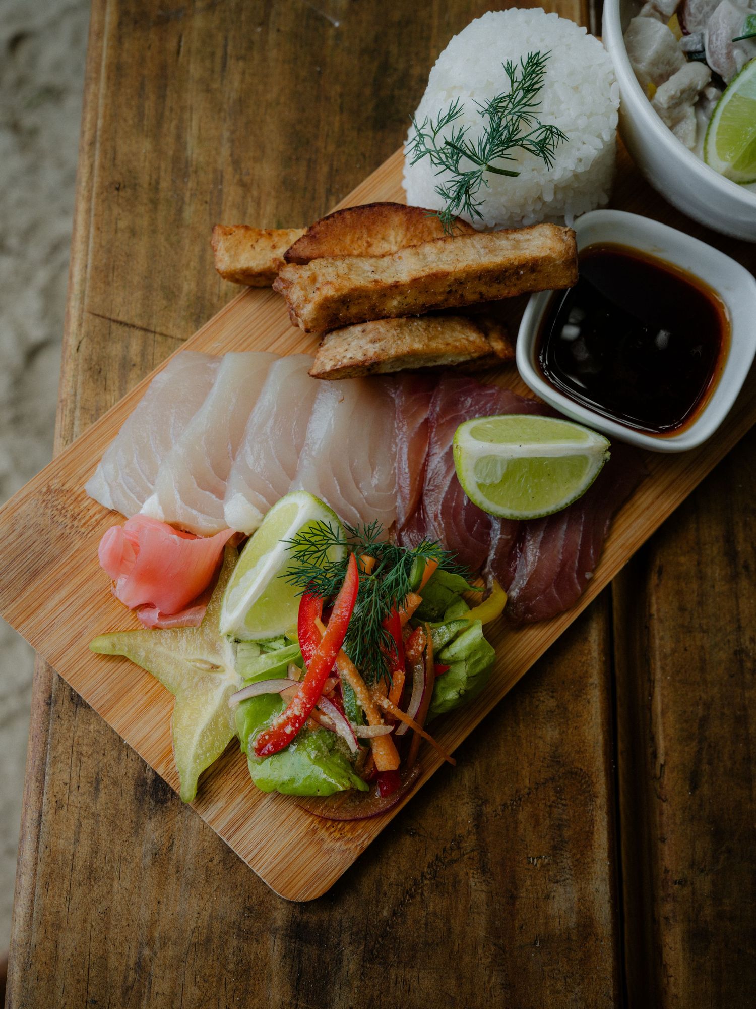 Sashimi platter on wooden board with soy sauce, lime, pickled ginger, salad, tofu, and rice garnished with dill.
