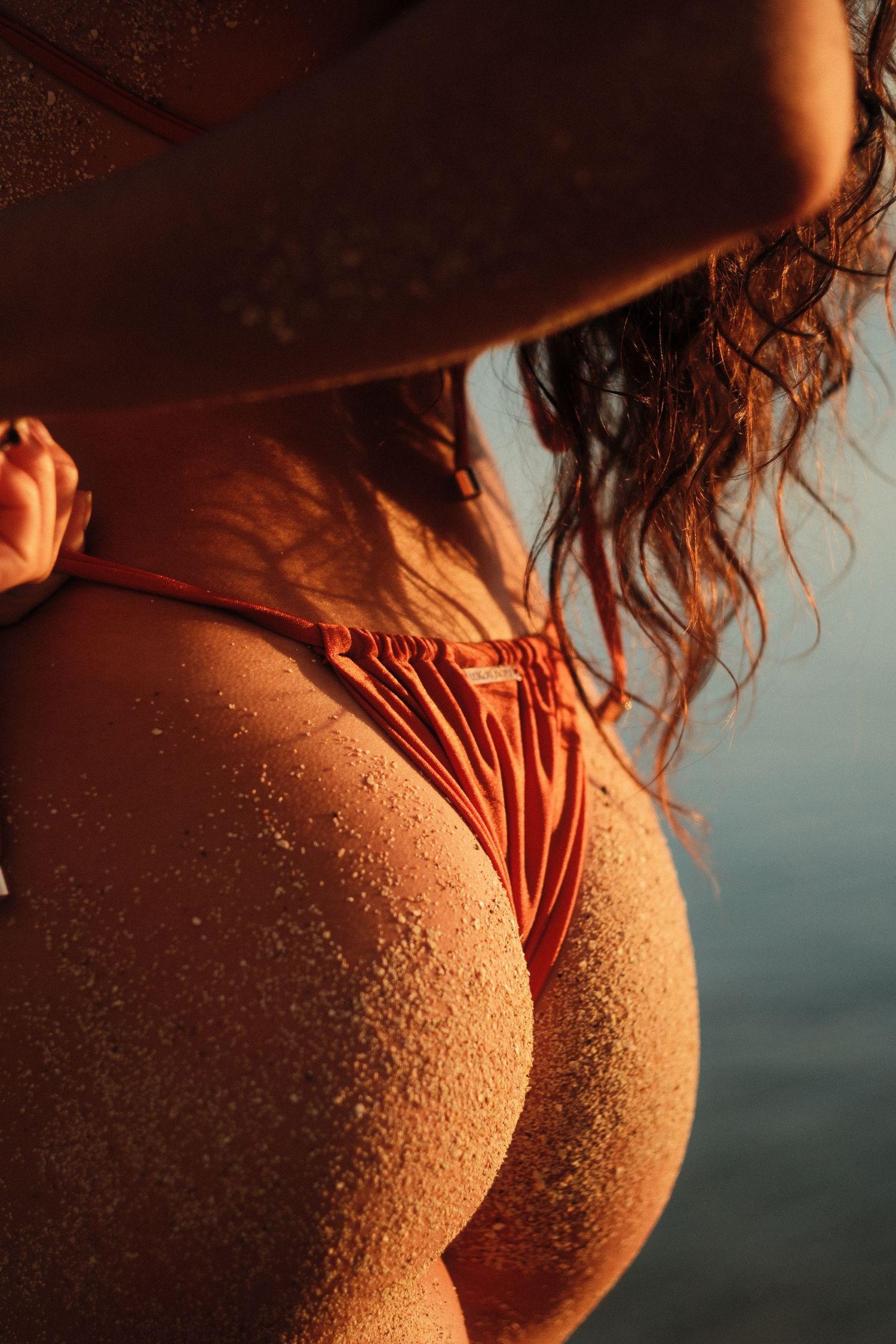 Woman in orange bikini on beach at sunset with sand-covered skin and ocean backdrop.